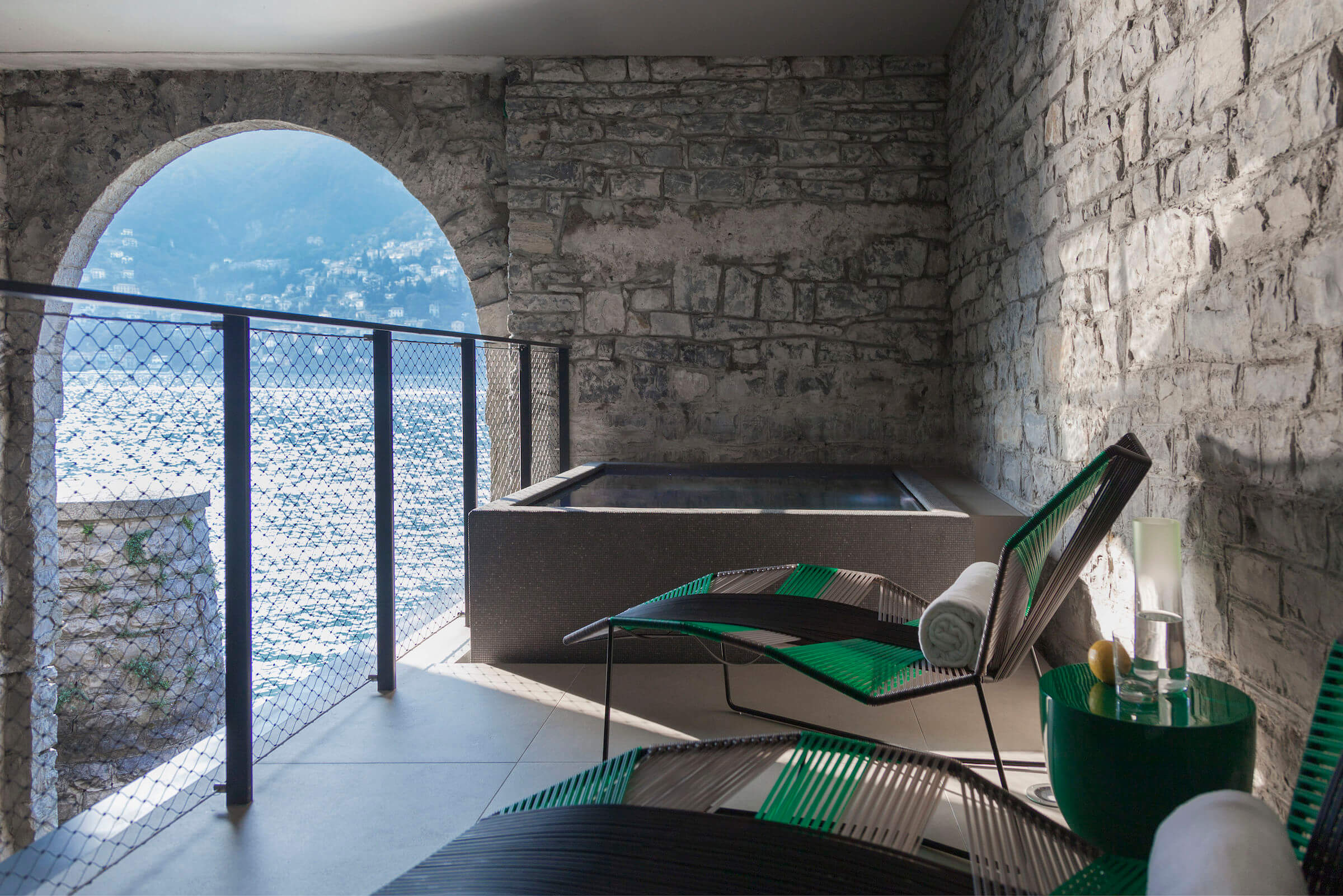 SPA at Il Sereno, Europe — Stone archway overlooking water, green woven lounge chairs, soaking tub, natural light through barrel vault opening.