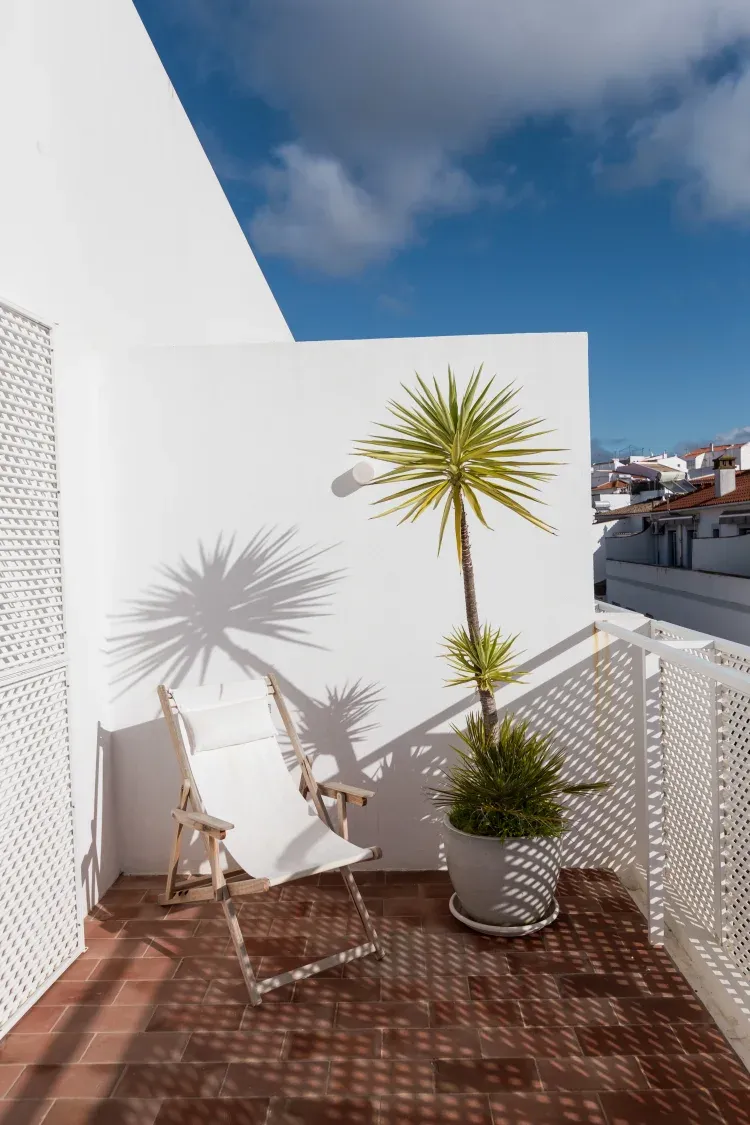 Casa Mãe, Europe — Minimalist rooftop terrace with white walls, wooden deck chairs, potted palms, geometric shadows, and blue sky framed by perforated screens.