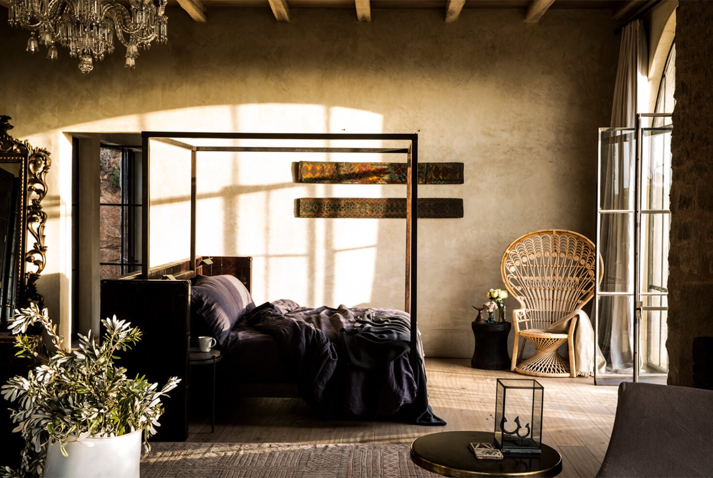 Arijiju Retreat, Africa — Luxury bedroom with black canopy bed, woven peacock chair, and golden light streaming through arched windows onto textured stone walls.