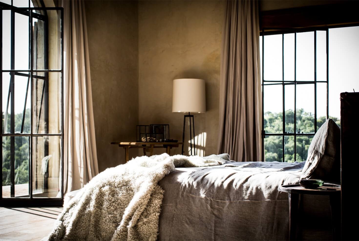 Arijiju Retreat, Africa — Minimalist bedroom with natural linen bedding, floor-to-ceiling windows, earthy plaster walls, black metal-frame doors, and expansive savanna views.