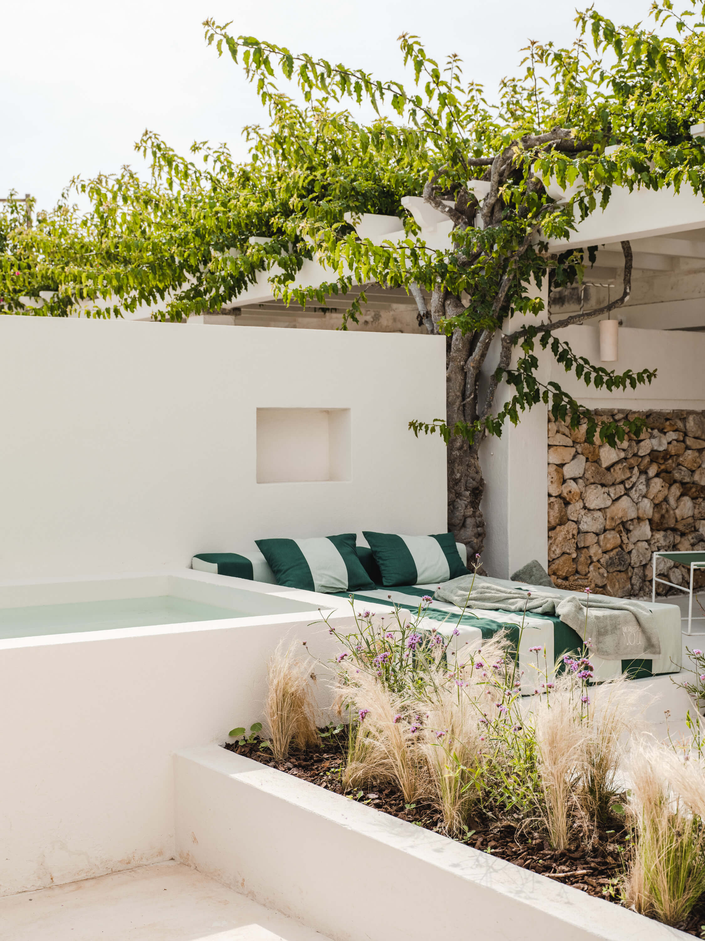 Morvedra Nou, Balearic Islands, Spain — minimalist poolside lounge with green striped cushions, ornamental grasses, mature tree, whitewashed architecture and stone wall detail.