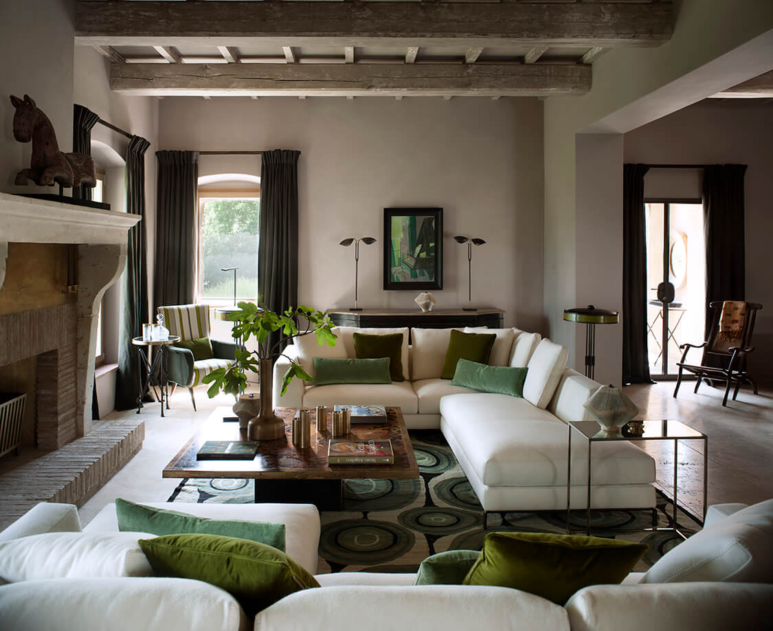 Castello di Reschio, Europe — Cream sectional sofa with green accents in bright living room with exposed beams, fireplace, arched windows, and geometric patterned rug.