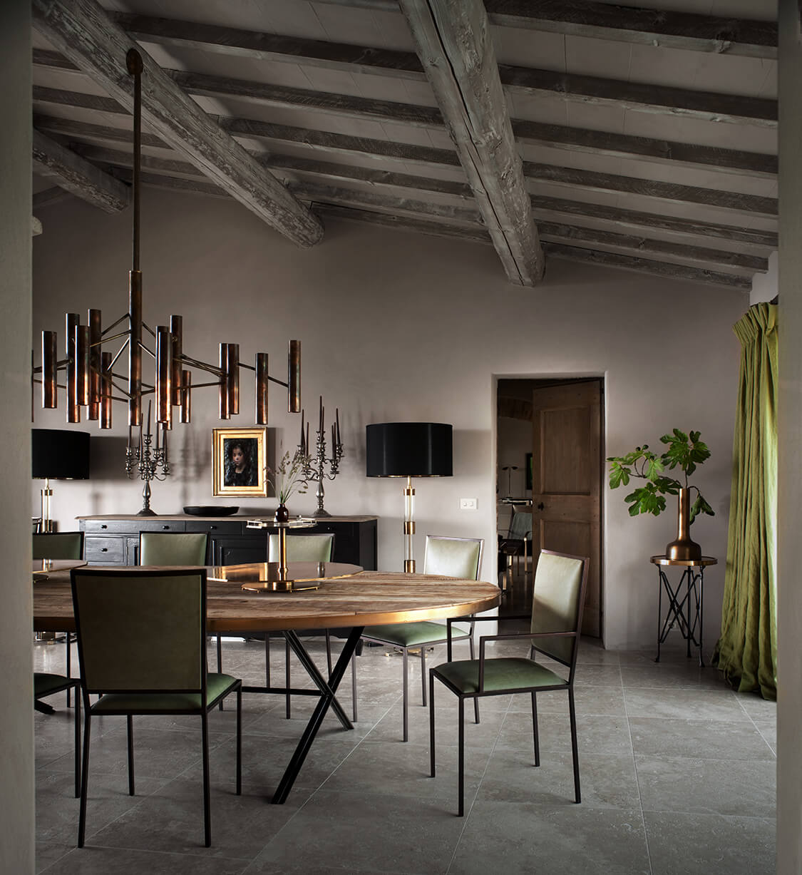 Castello di Reschio, Europe — Dining room with round timber table, black metal chairs, exposed beam ceiling, portrait artwork, and modern brass chandelier in historic stone interior.