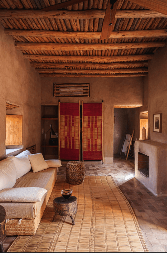Berber Lodge, Africa — Ochre-walled bedroom with exposed wooden beam ceiling, terracotta tile floors, cream upholstered daybed, and red textile room divider.