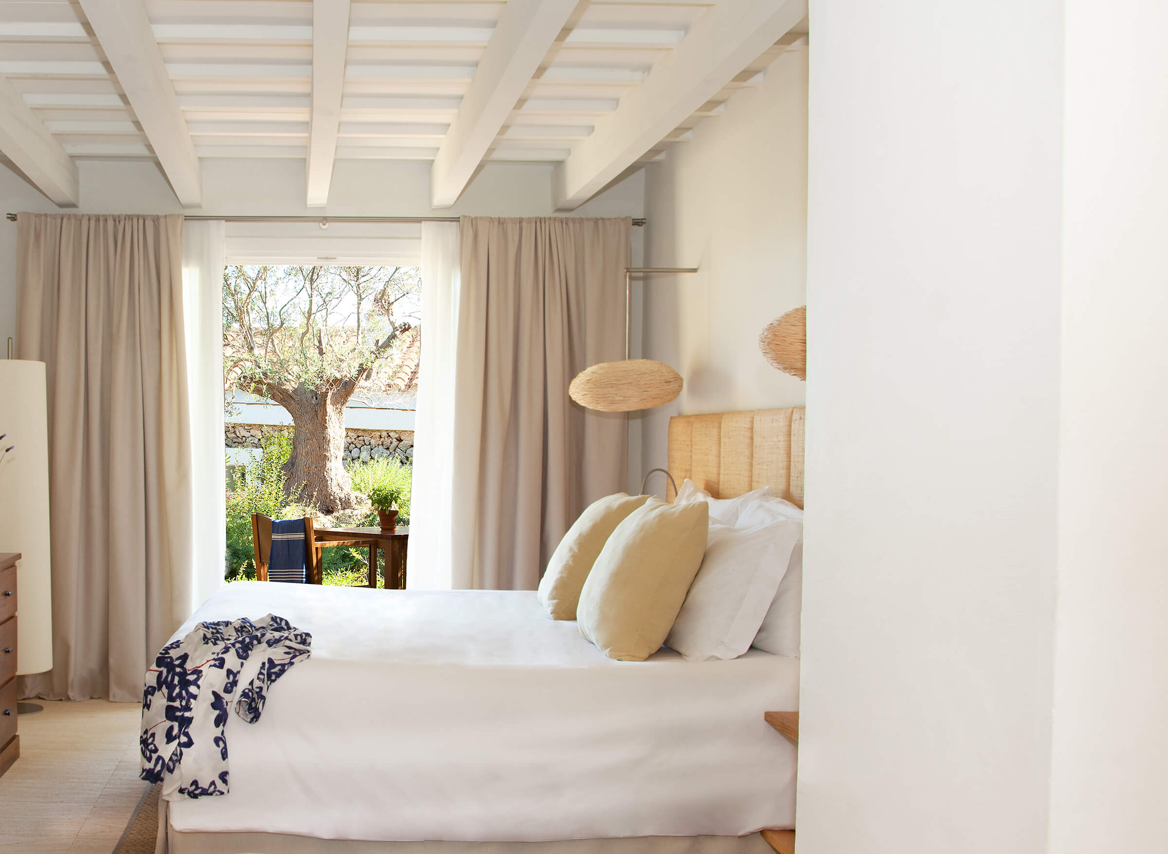 Torralbenc, Balearic Islands — Bedroom with woven headboard, white linens, exposed beam ceiling, and terrace view of ancient olive tree.