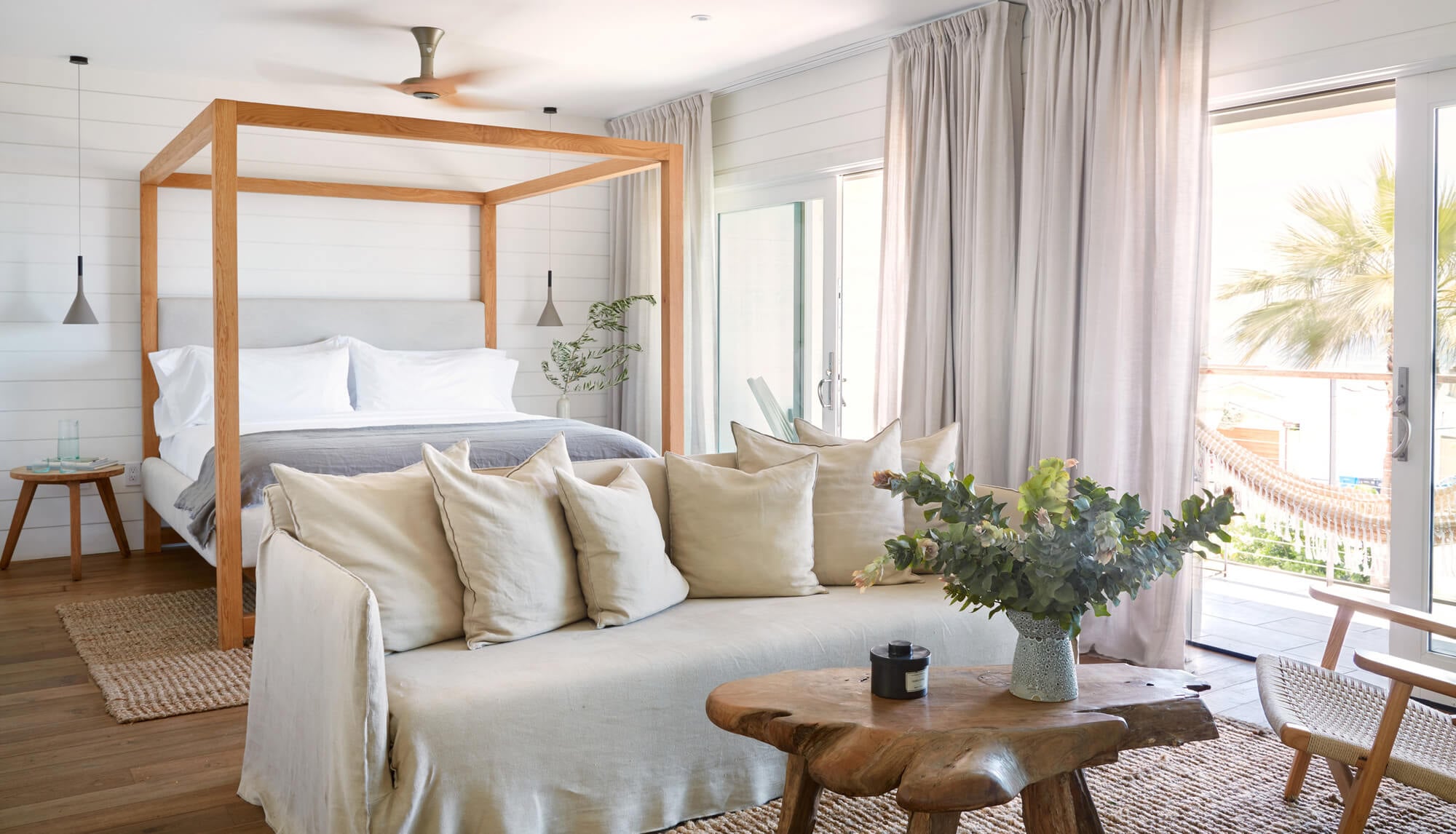 The Surfrider, North America — Coastal bedroom with wooden four-poster bed, cream linen sofa, natural light from ocean-view terrace, shiplap walls, and warm wood furnishings.