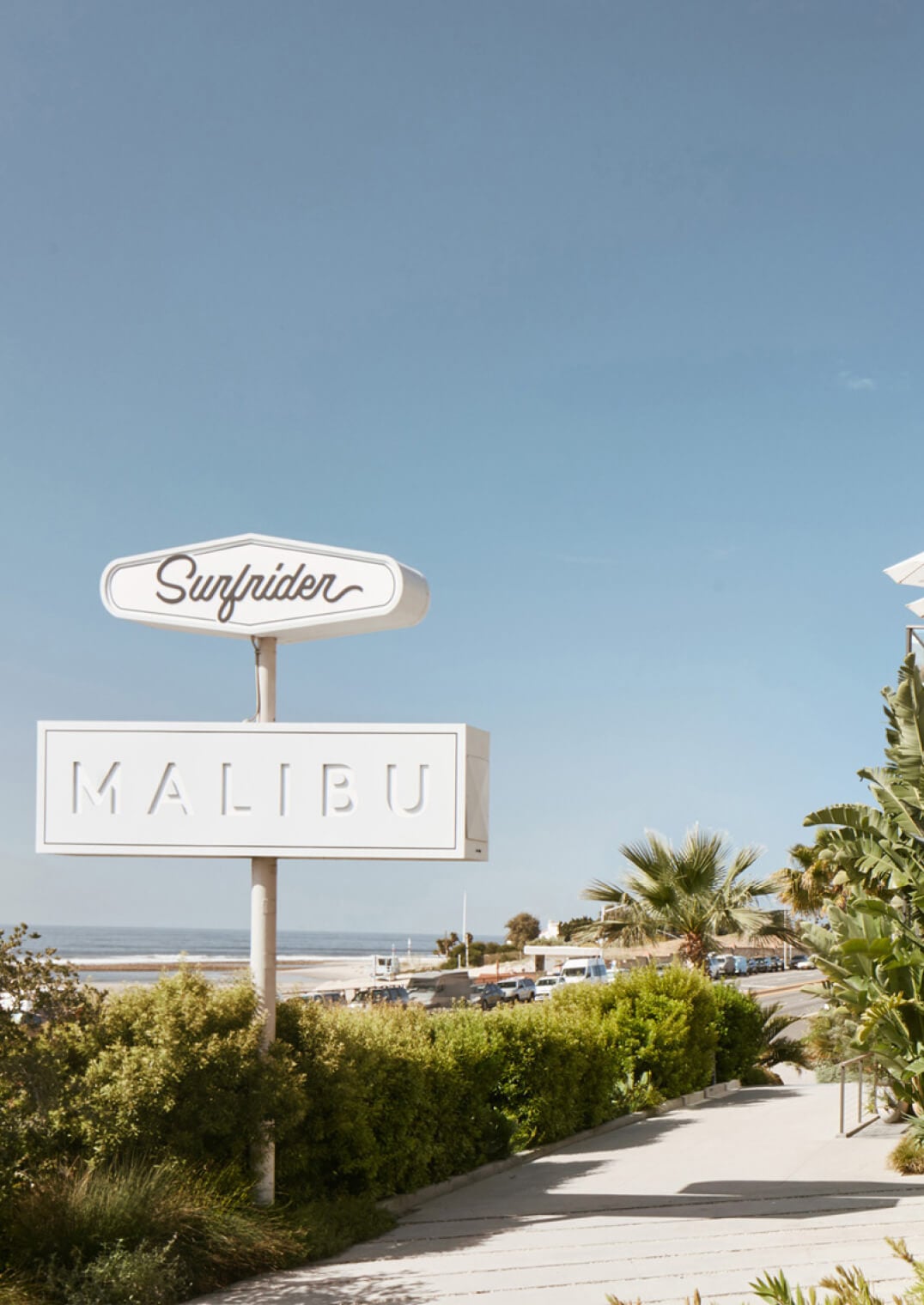 The Surfrider, Malibu, USA — Vintage neon sign with ocean view, palm trees, and coastal architecture under clear blue sky.