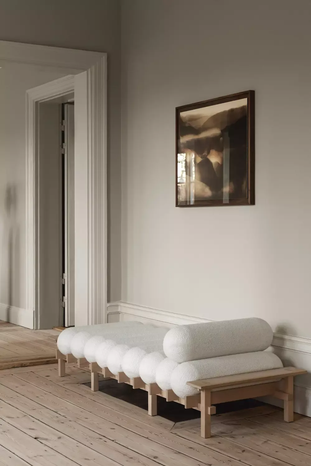 House of Gärsnäs, Europe — Minimalist wooden bench with white upholstered cylindrical cushions in bright hallway with light wood floors and cream walls.