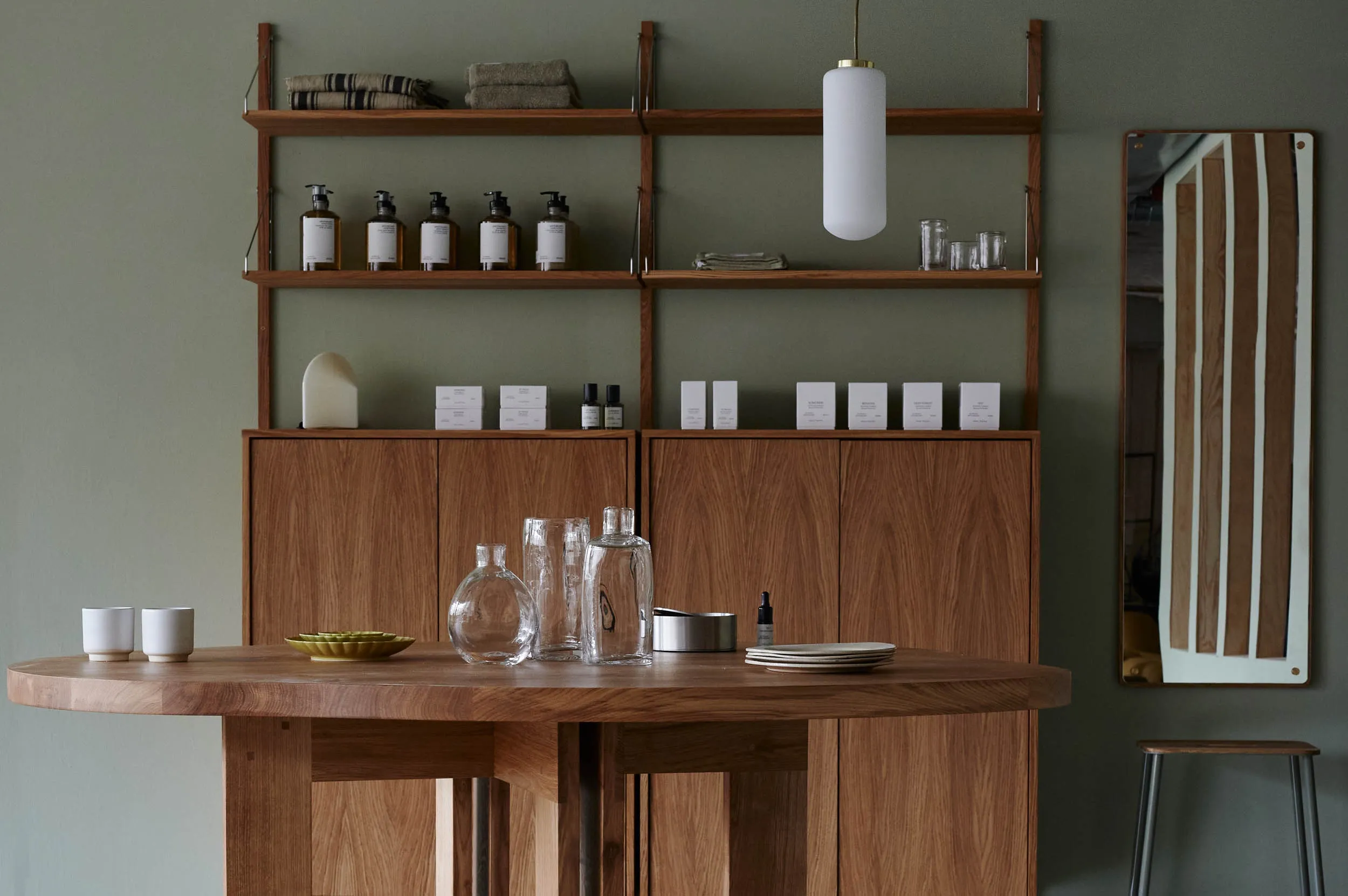 Tambur, Europe — Walnut shelving system with ceramic vessels, glass bottles, and white bath products against sage green wall, minimalist hospitality design.