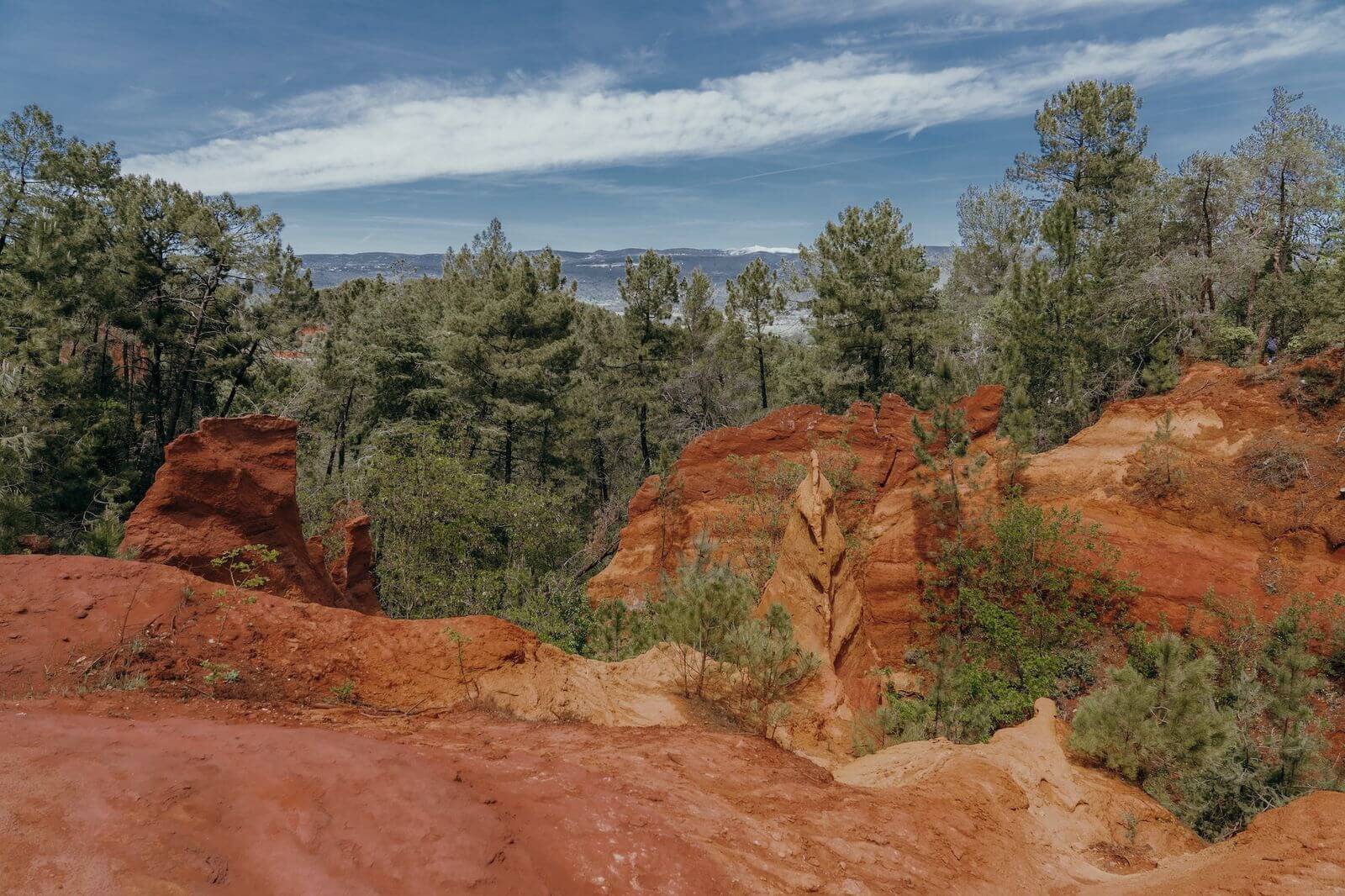 The Ocher Trail in Roussillon — Ocher Trail, Roussillon, France — rust-red mineral cliffs and pine forest overlooking distant valley under blue sky, natural pigment landscape