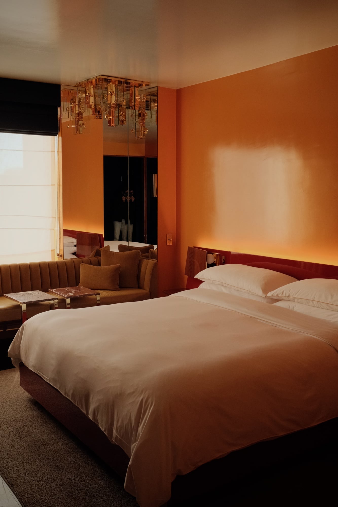 The Manner, North America — Warm ochre bedroom with brass pendant lighting, cream bedding, cognac leather seating, and floor-to-ceiling mirror reflecting ambient golden light.