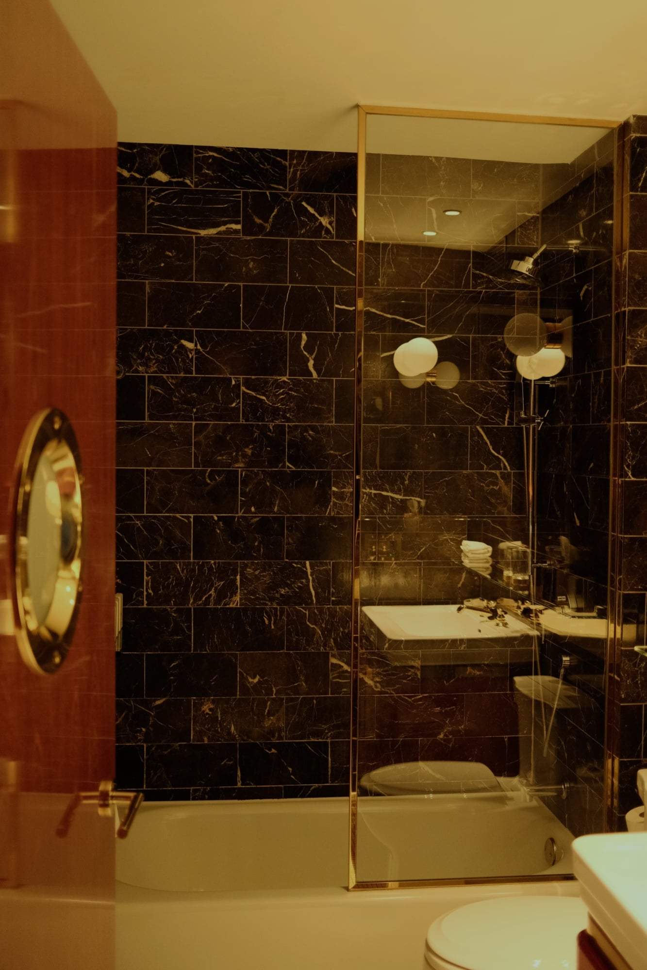 The Manner, North America — Luxury bathroom with black marble tile walls, brass-framed glass shower enclosure, and warm ambient lighting reflecting off polished surfaces.
