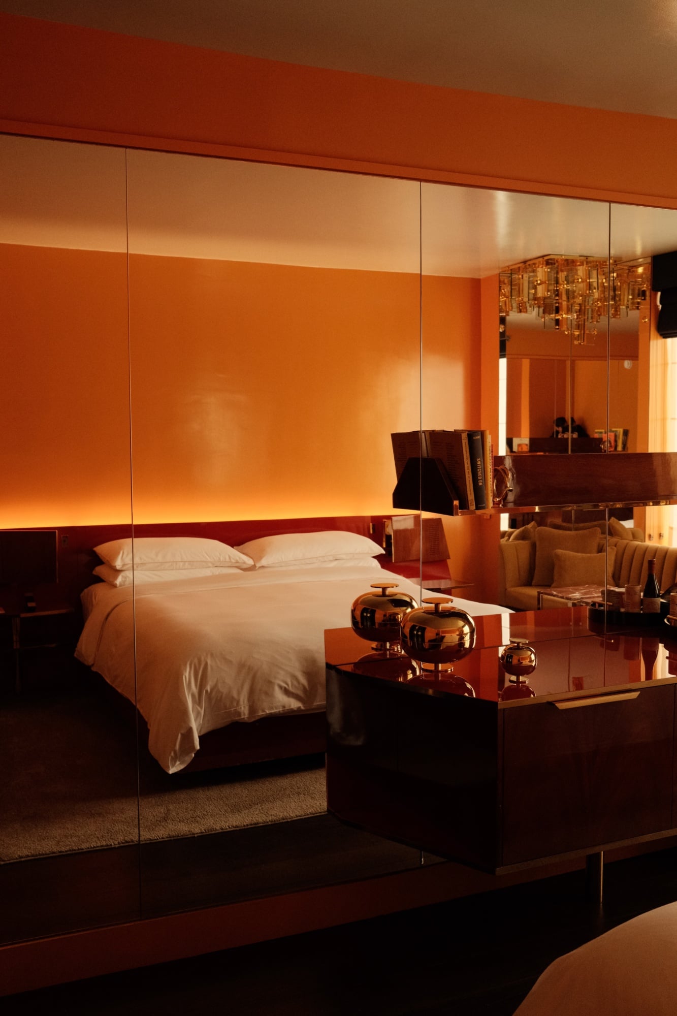 The Manner, North America — warm-lit modern hotel bedroom with mirrored glass partition, copper accents, white linens, and sculptural metallic decor on dark wood surfaces.