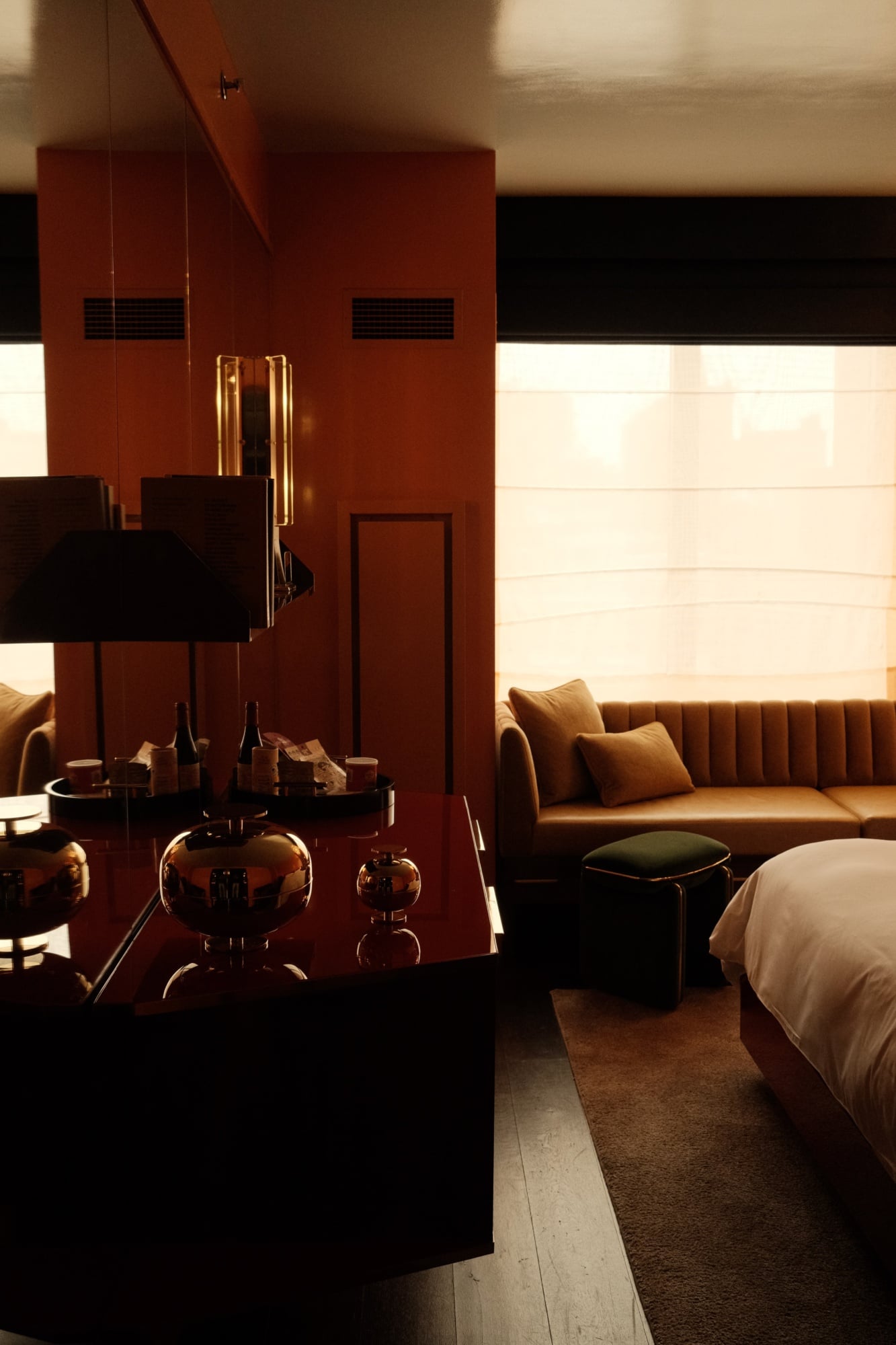 The Manner, North America — Warm ochre sofa with black accent pillow beside bed in brutalist hotel room with terracotta partition wall and golden hour sunlight through floor-to-ceiling windows.