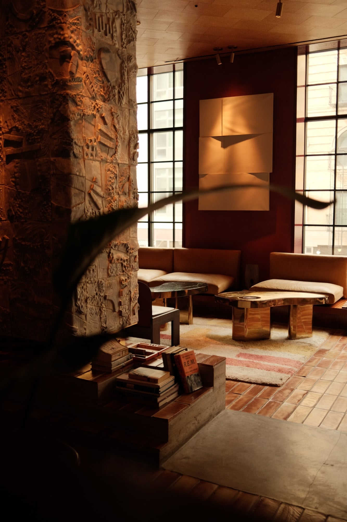 The Apartment, North America — Warm minimalist lounge with carved stone column, terracotta tile flooring, leather seating, and gridded windows casting geometric light across ochre walls.