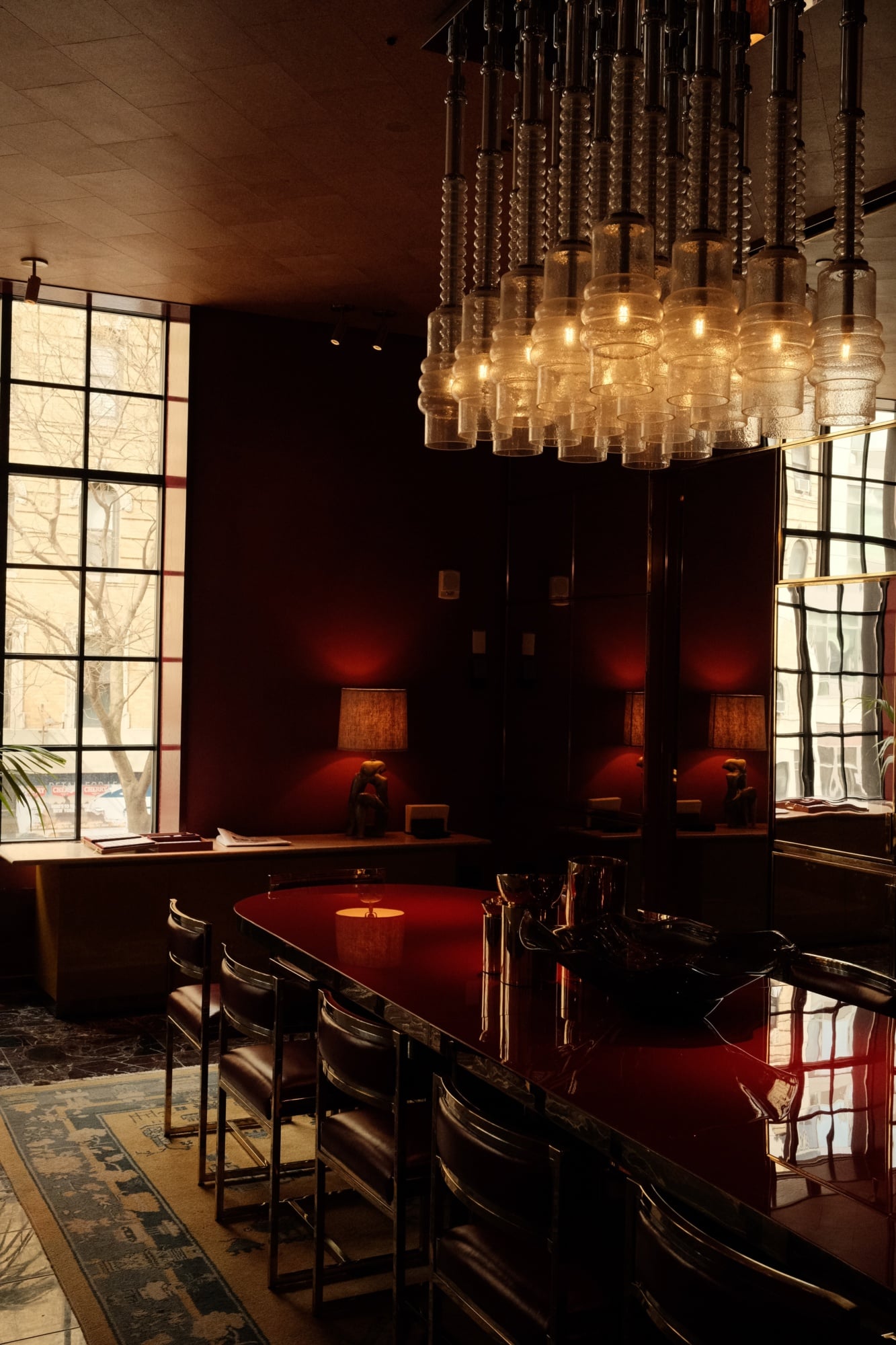 The Manner, North America — Luxury hospitality lounge with burgundy lacquered grand piano, suspended glass pendant chandelier, dark wood paneling, gridded windows, and warm ambient lighting.