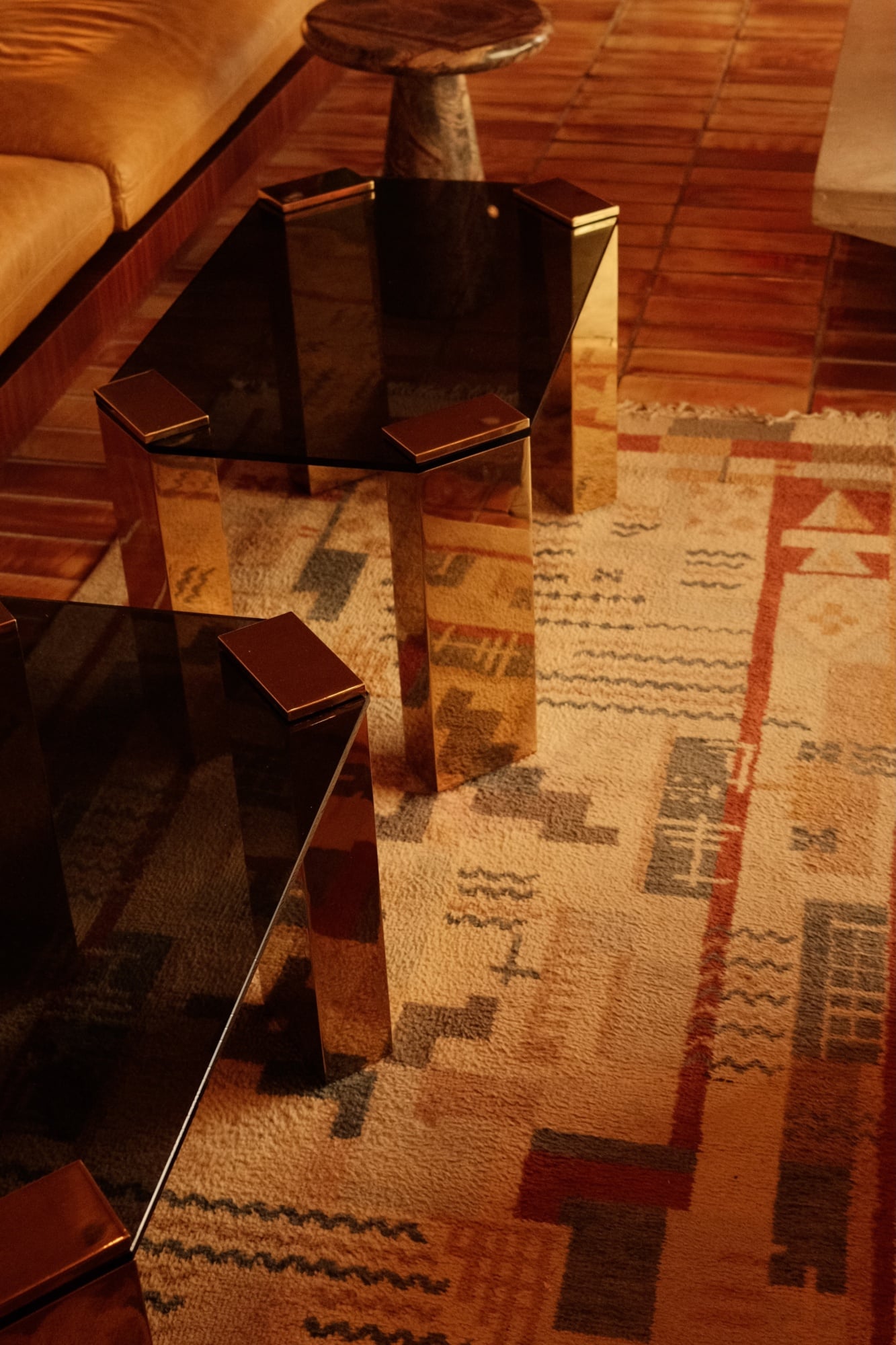The Manner, North America — Modern glass coffee table with wooden legs casting shadows on geometric patterned rug, warm wood paneling and natural light.