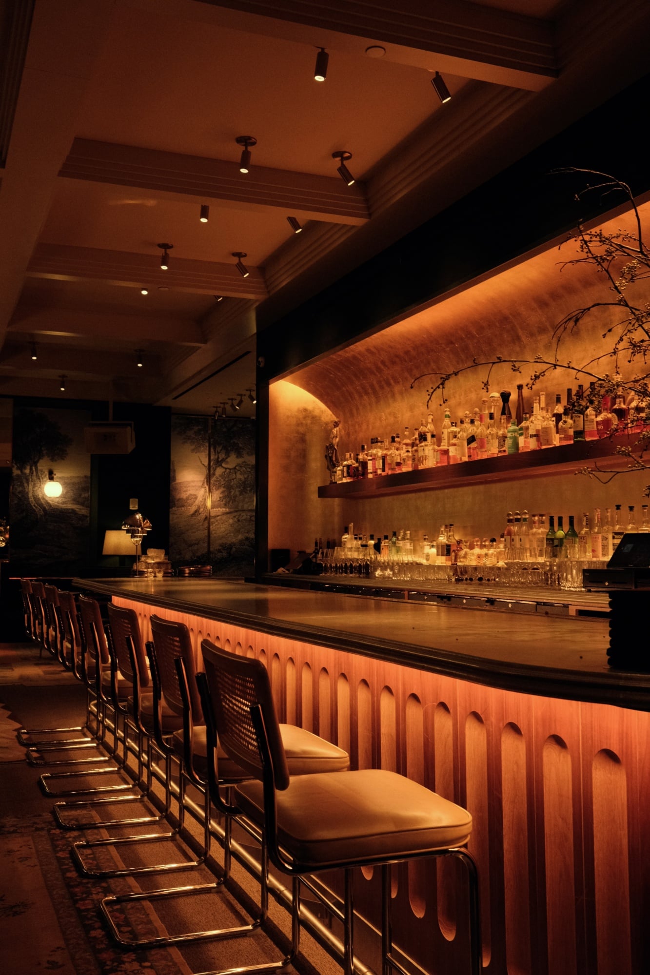 Sloane's, North America — Warm-lit bar with curved wood paneling, backlit spirit shelves, coffered ceiling, and brass bar seating overlooking moody textured walls and ambient lighting.