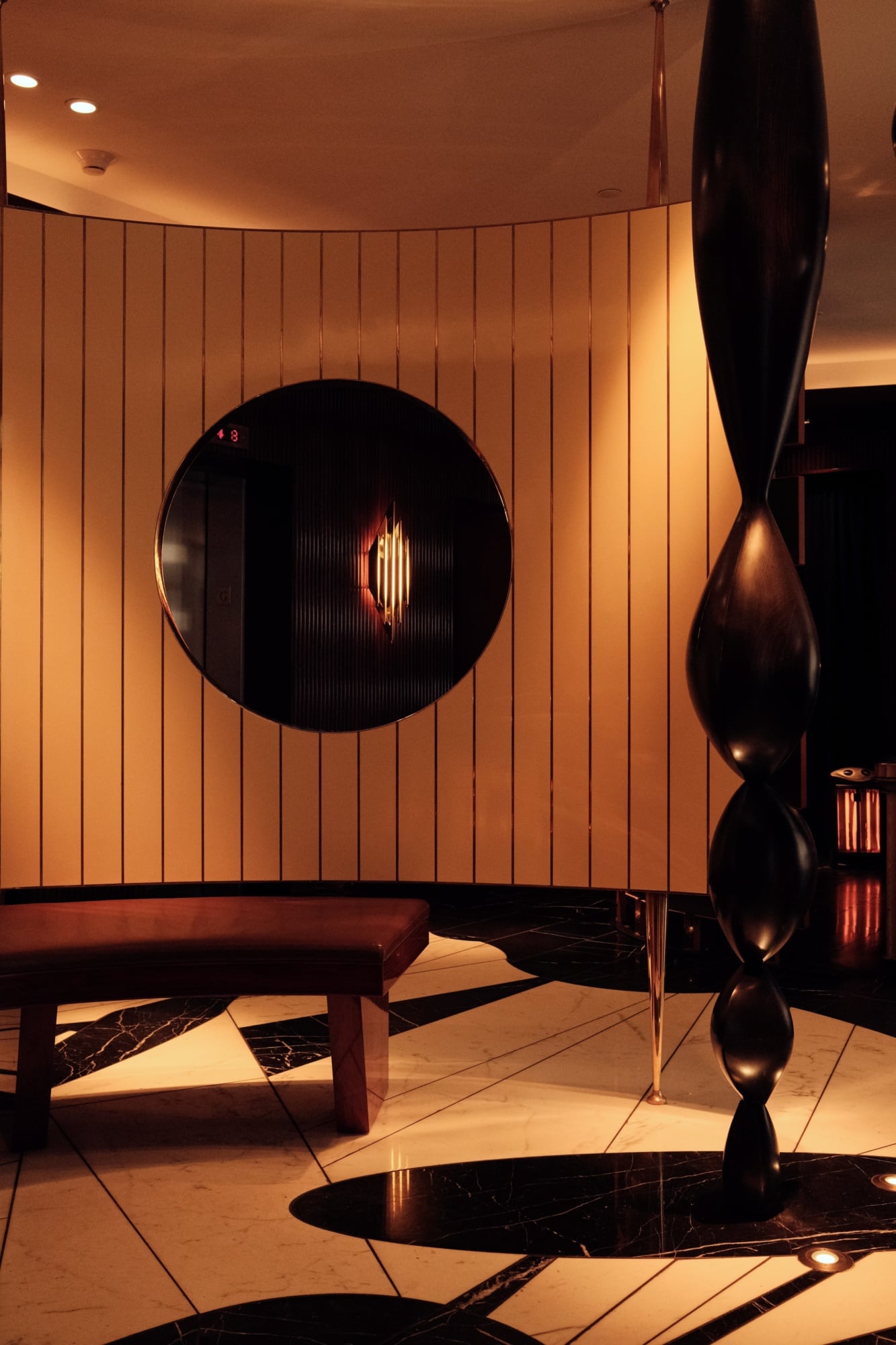 The Manner, North America — Minimalist lounge with warm wood-paneled walls, circular window portal, sculptural black forms, and marble flooring bathed in ambient golden lighting.