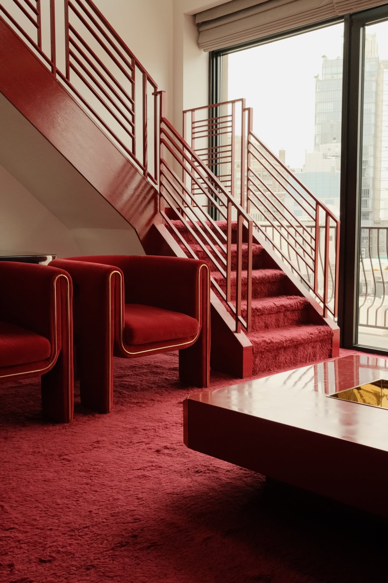 The Manner, North America — Burgundy velvet armchair with brass piping beside geometric staircase, natural light from floor-to-ceiling windows overlooking cityscape.