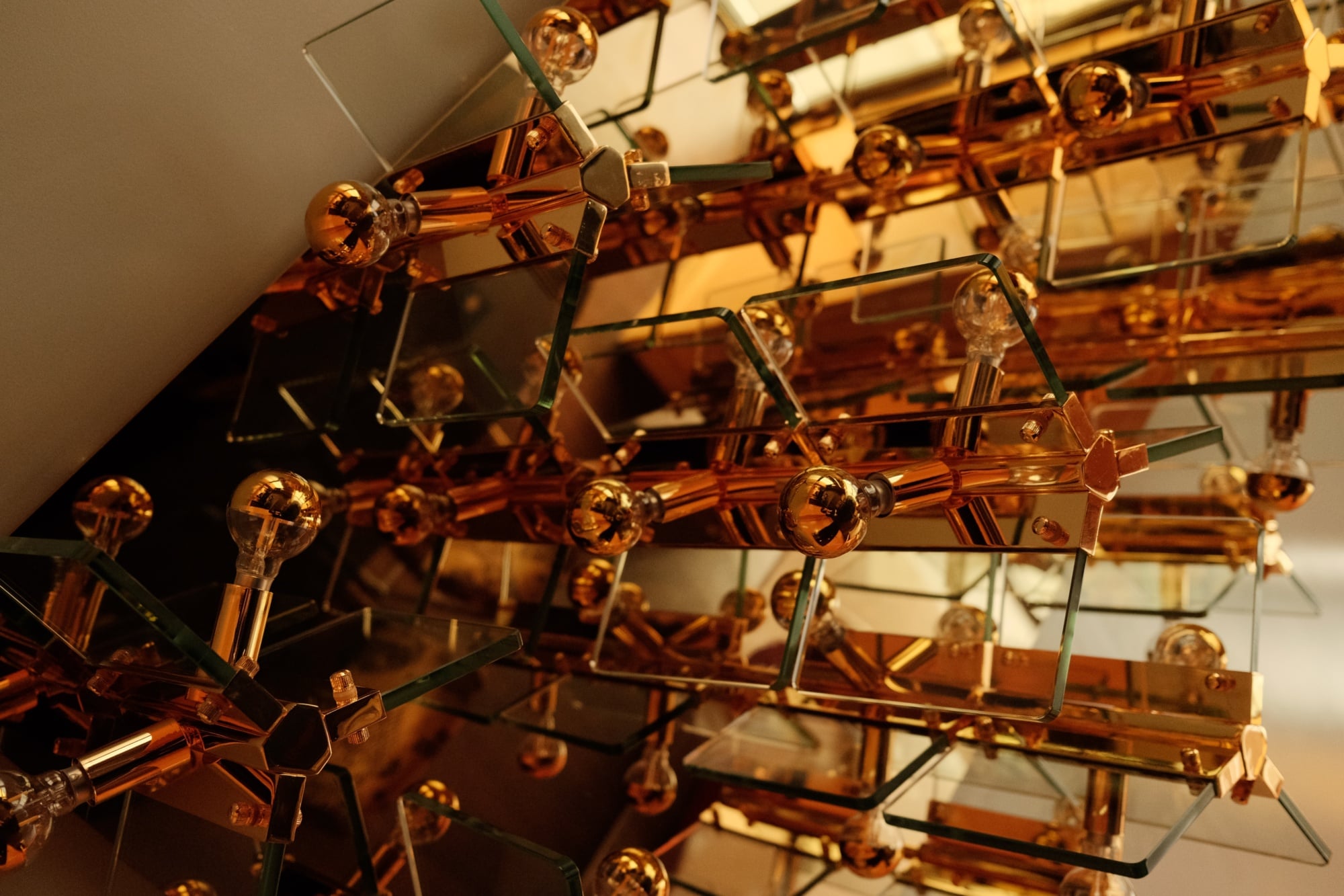 The Manner, North America — Amber glass sculptural installation with spherical finials and geometric brass framework, casting warm light and reflections on white wall.