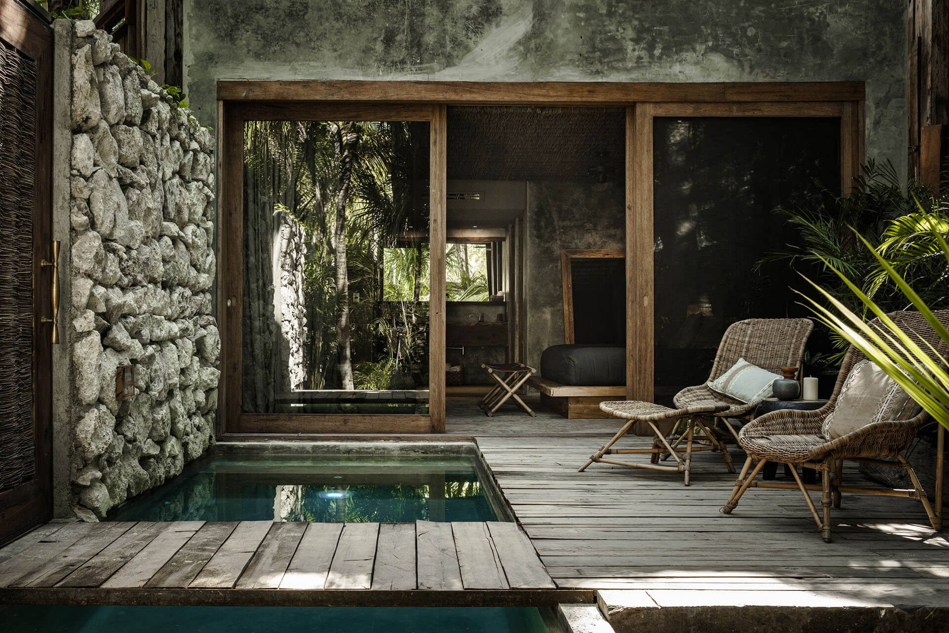 Be Tulum, Mexico — Wooden deck terrace with sunken plunge pool, woven rattan chairs, concrete walls, and glass doors opening to jungle vegetation and bedroom interior.