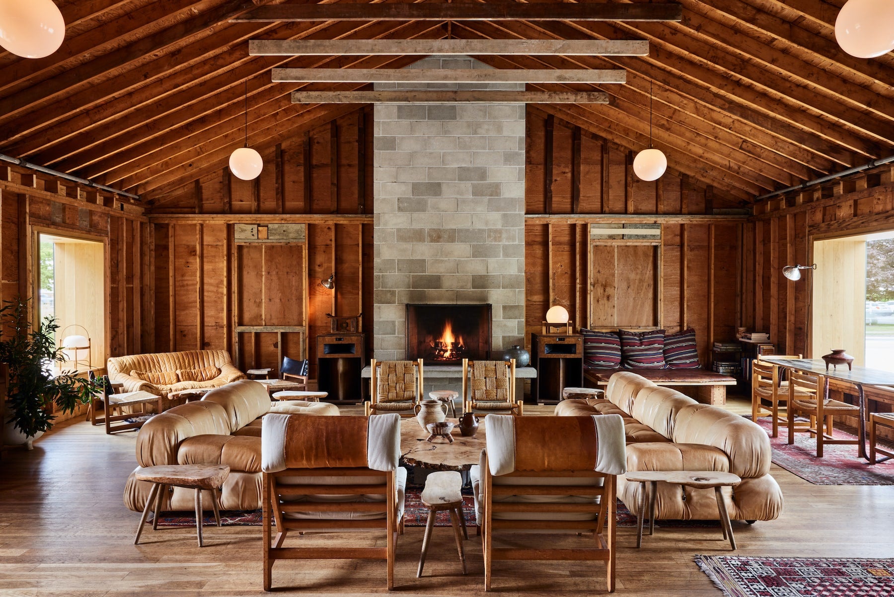 Tourists, North America — Timber-framed lodge interior with central stone fireplace, cognac leather seating, exposed beams, and warm ambient lighting.