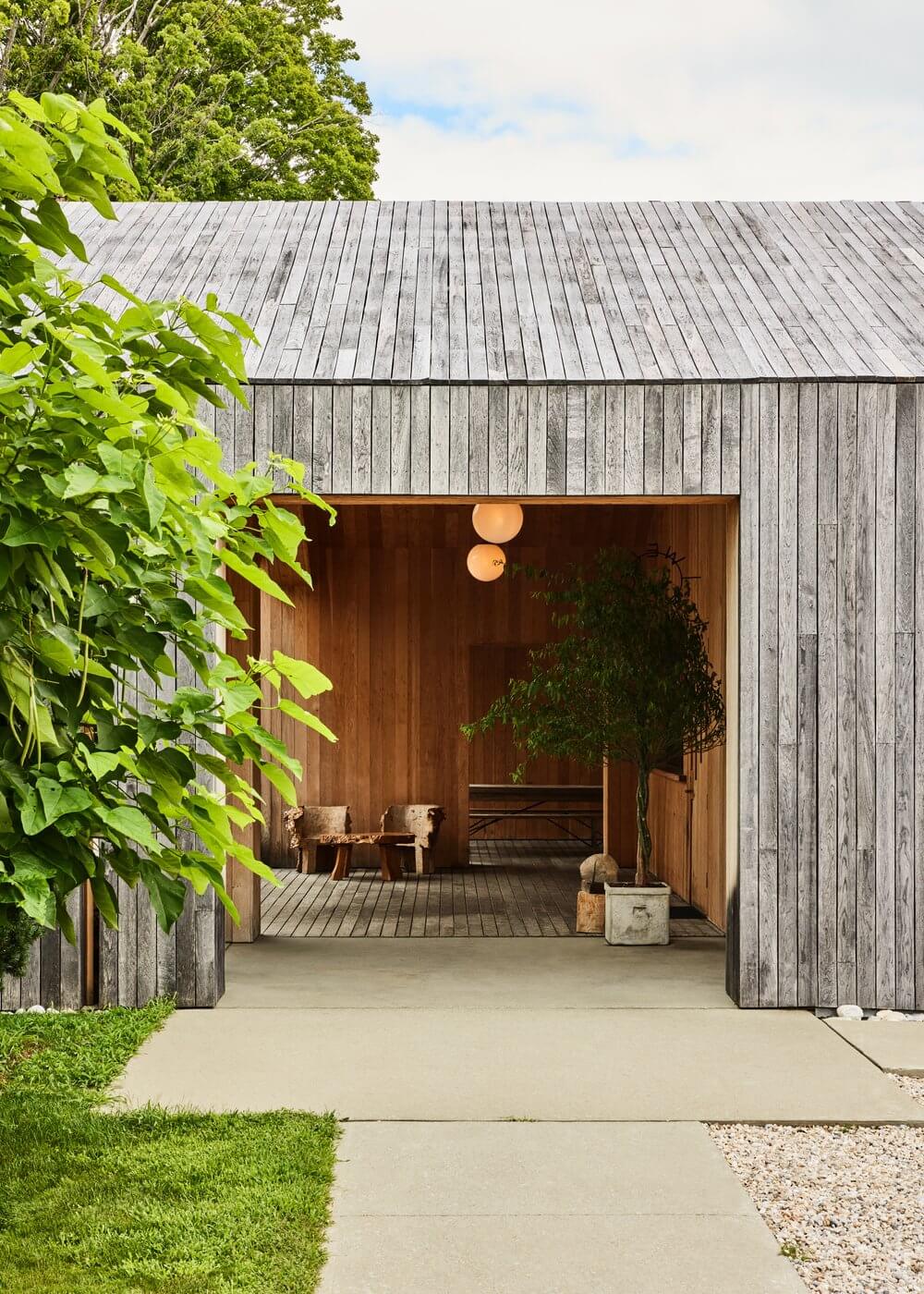Tourists, North America — Weathered timber pavilion with warm wood interior, pendant lights, and open deck overlooking manicured grounds, minimalist modern architecture.