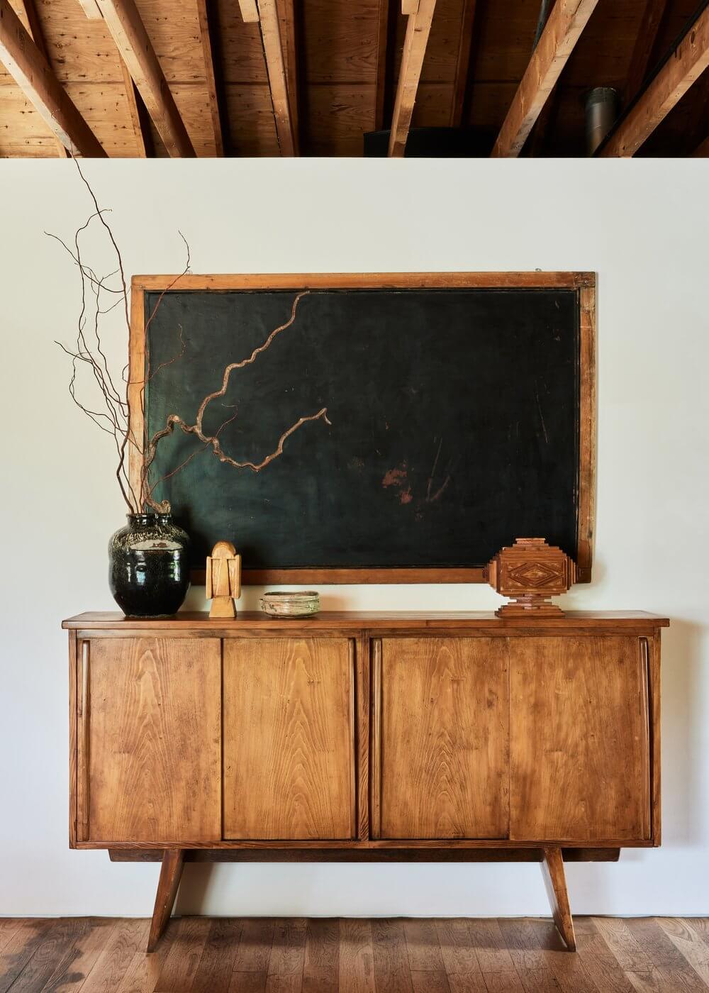 Tourists, North America — Mid-century wooden sideboard with black chalkboard and brass accents beneath exposed beam ceiling, minimalist hospitality interior design.