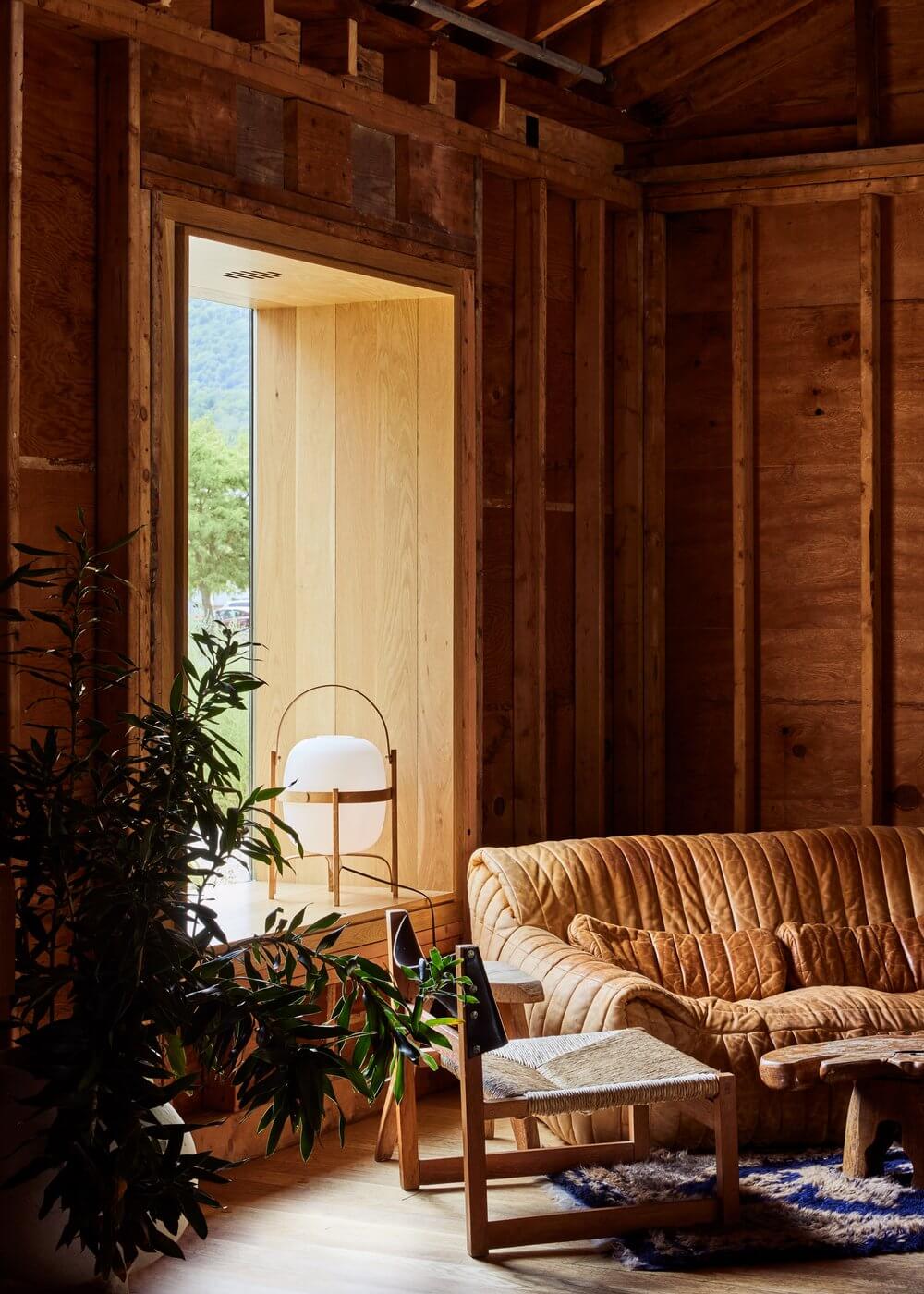 Tourists, North America — Wooden lodge interior with tan leather sofa, woven chair, potted plant, and floor lamp overlooking mountain landscape through glass doors.
