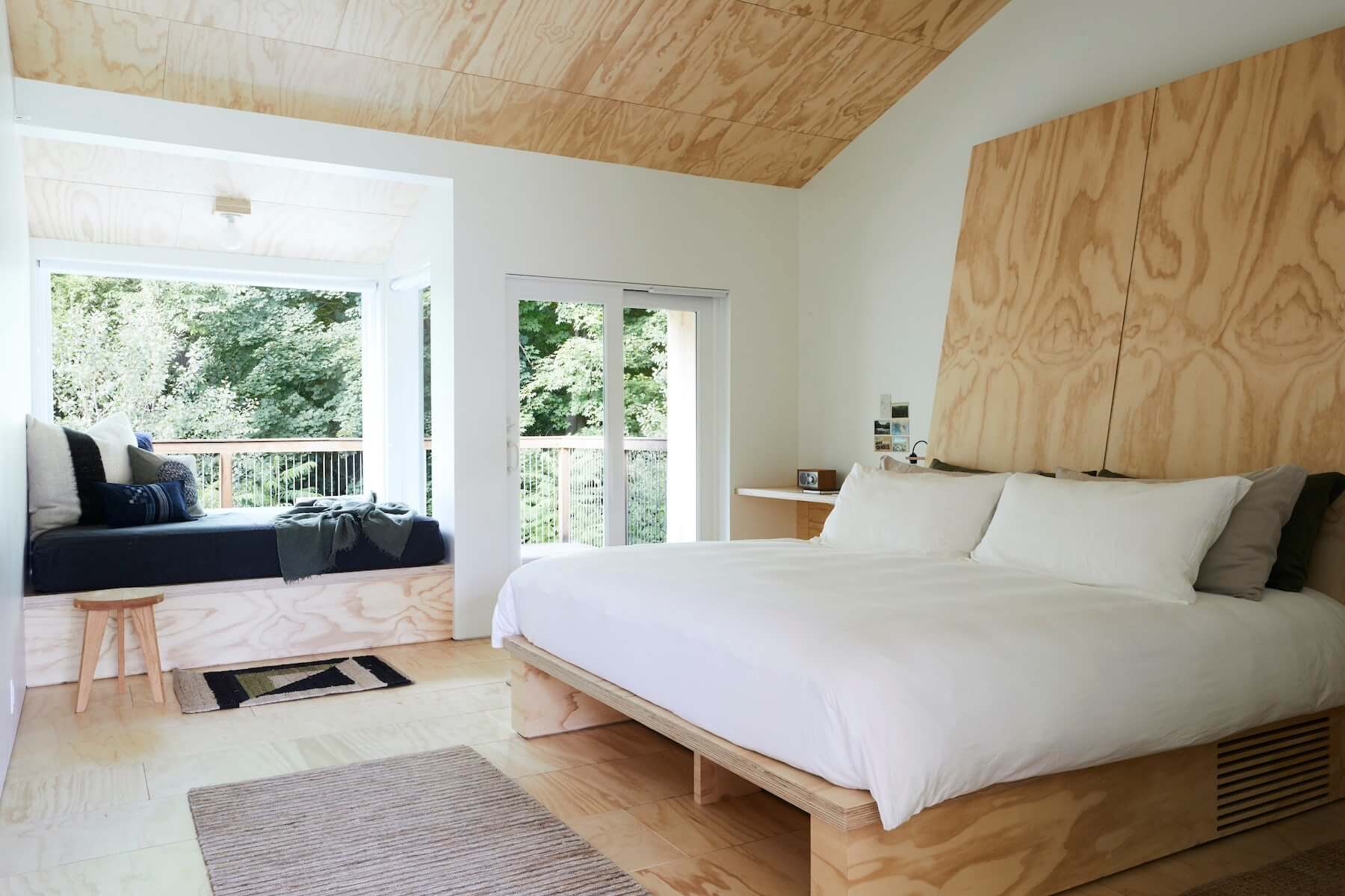 Tourists, North America — Minimalist bedroom with plywood ceiling, natural wood bed frame, white linens, large window seat overlooking forest, scandinavian modern design.