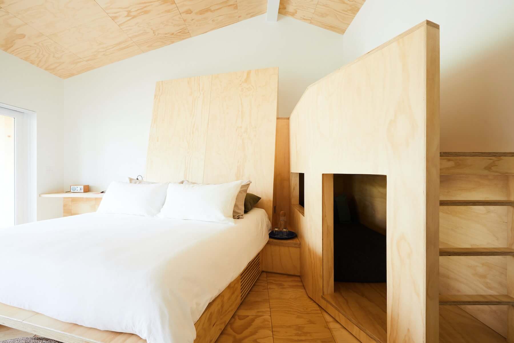 Tourists, North America — Minimalist plywood bedroom with curved wooden partition, built-in bed with white linens, skylights, and warm natural light throughout bright attic space.