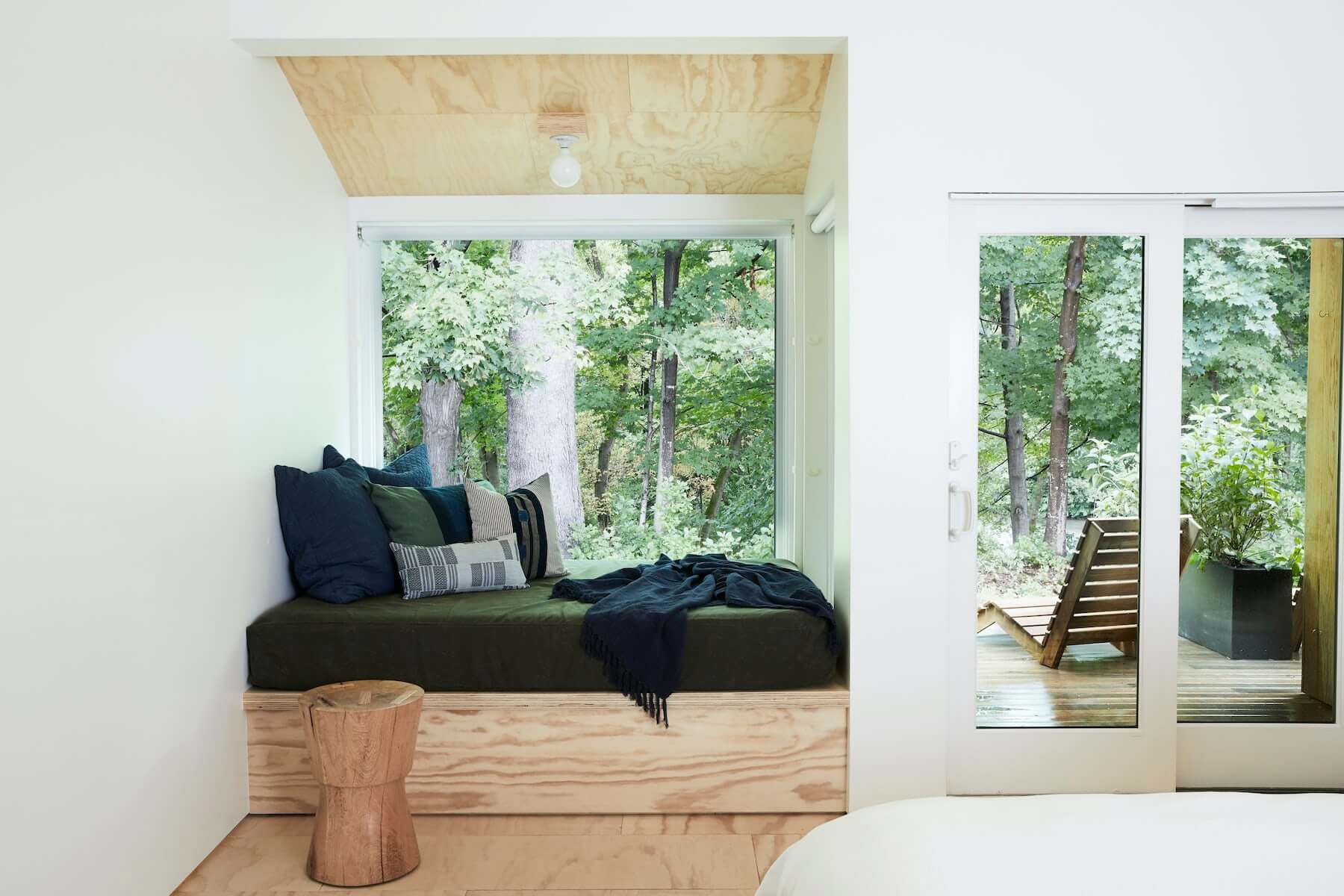 Tourists, North America — Modern cabin bedroom with built-in window seat, dark upholstery, forest views, plywood ceiling, wooden stool, glass doors to deck.
