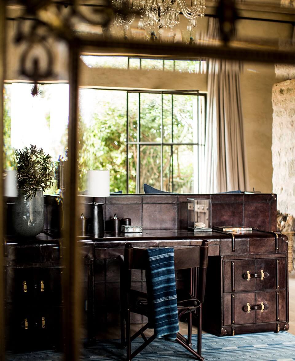 Arijiju Retreat, Africa — Dark leather desk with brass hardware, blue striped textile, crystal chandelier, floor-to-ceiling windows overlooking lush greenery, minimalist luxury study.