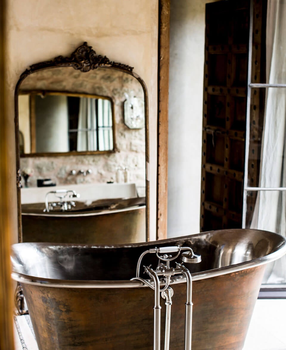 Arijiju Retreat, Africa — Vintage copper bathtub with polished brass fixtures and ornate black mirror reflecting minimalist bathroom in cream stone villa.