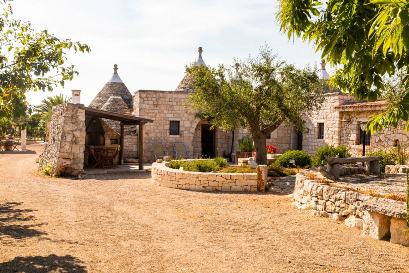 Trulli Il Castagno Winery, Italy — Conical-roofed trulli stone buildings with terracotta finials, courtyard garden, dappled sunlight, Mediterranean architecture.