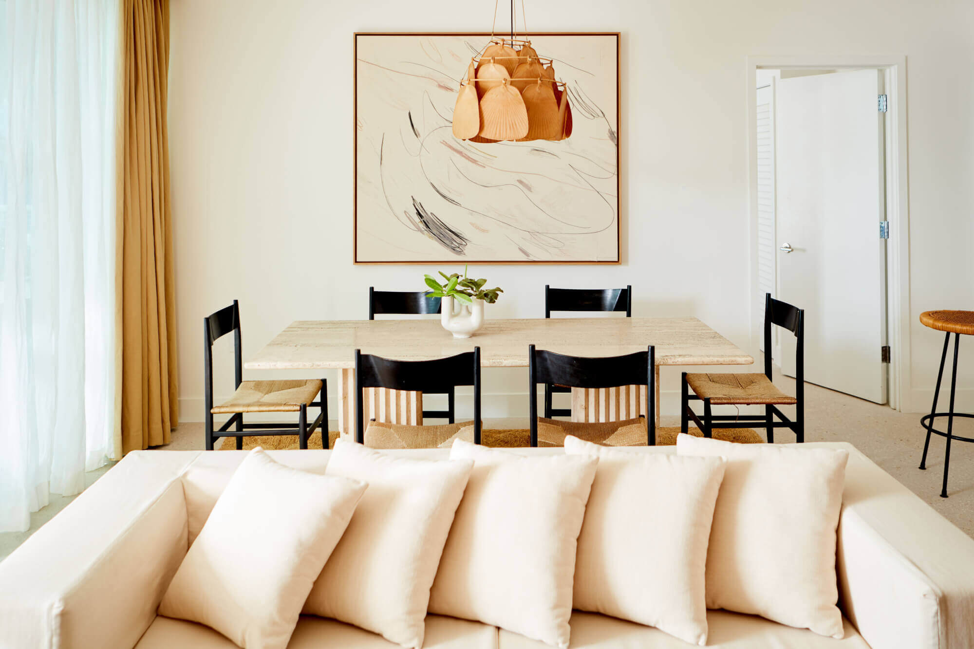 Palm Heights, North America — Minimalist dining room with light oak table, black chairs, woven rush seating, cream sofa with pillows, abstract artwork, pendant lighting, and golden curtains.