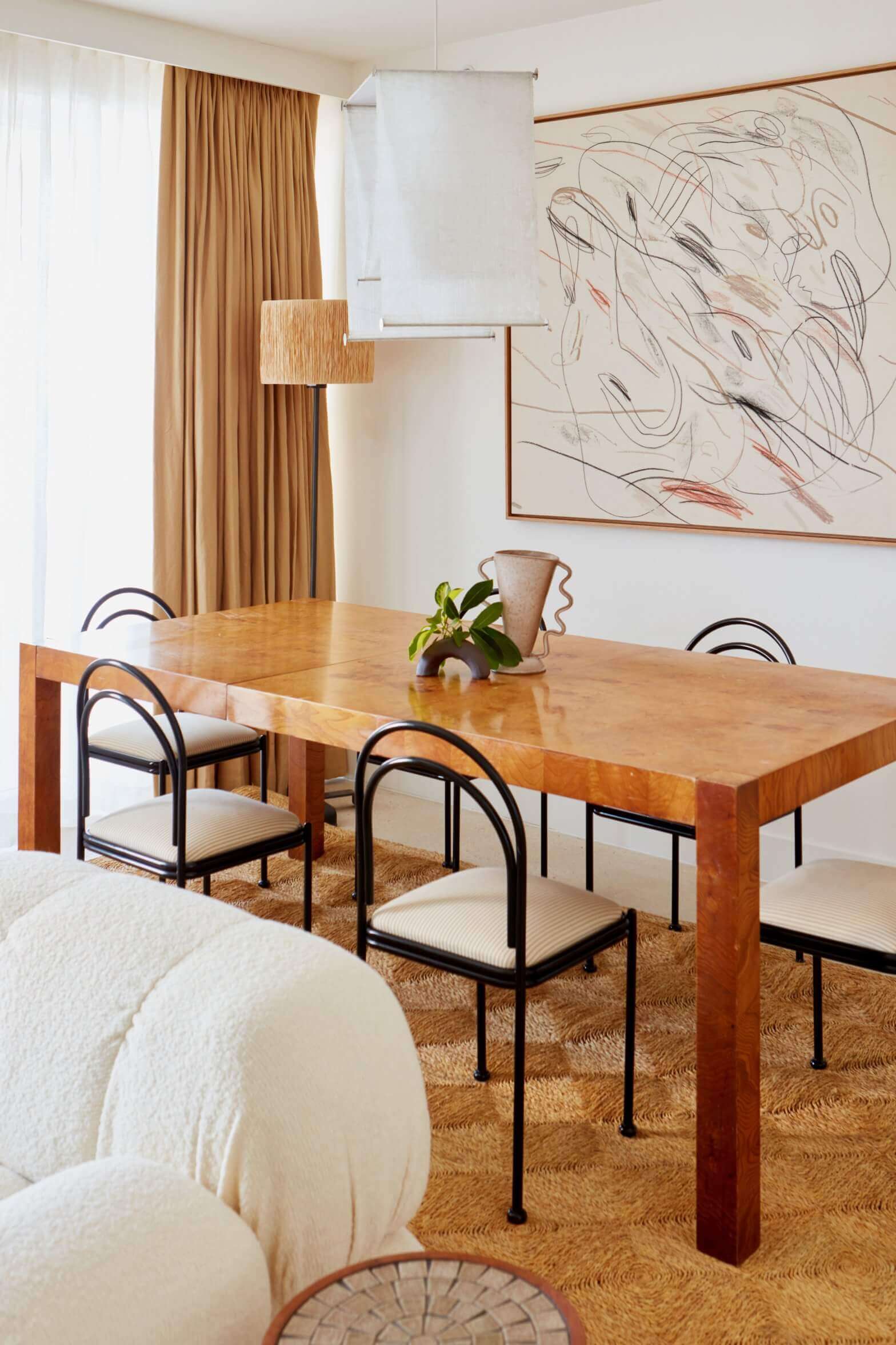 Palm Heights, North America — Modern dining room with warm wood table, curved black metal chairs, terracotta curtains, abstract wall art, and natural light from floor-to-ceiling windows.