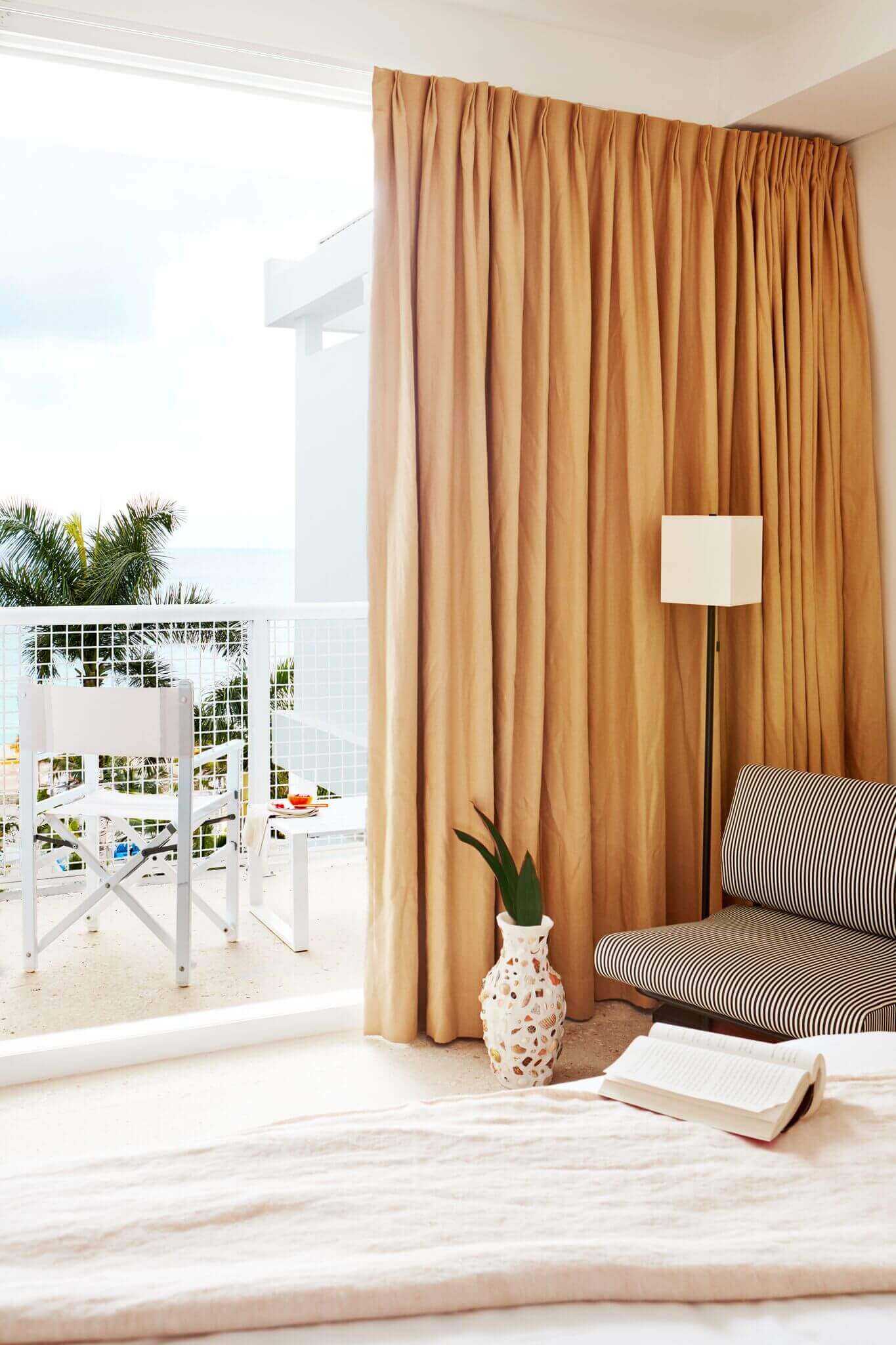 Palm Heights, North America — Minimalist coastal bedroom with golden curtains, striped daybed, terracotta vase, open balcony overlooking ocean and palm trees.
