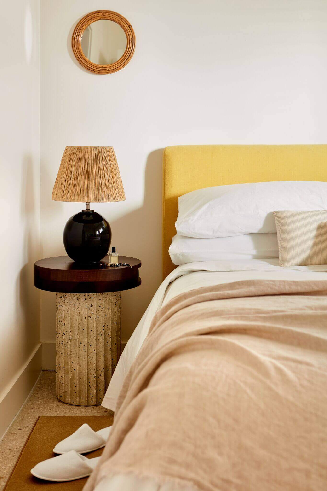 Palm Heights, North America — Minimalist bedroom corner with mustard upholstered headboard, cork pedestal nightstand, black ceramic lamp, woven mirror, and neutral linens in natural light.