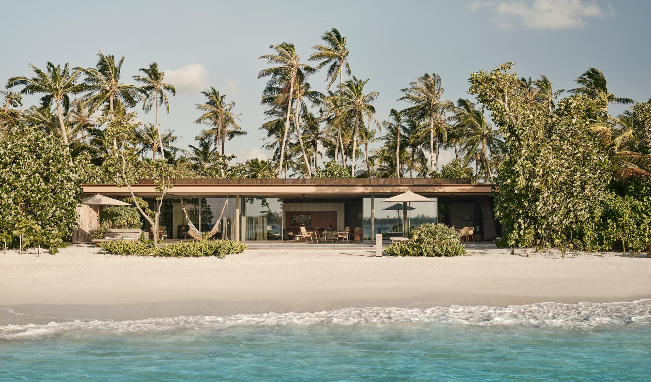 Patina Maldives — Modernist beachfront villa with horizontal timber roof, floor-to-ceiling glazing, surrounded by coconut palms and lush vegetation on white sand beach.