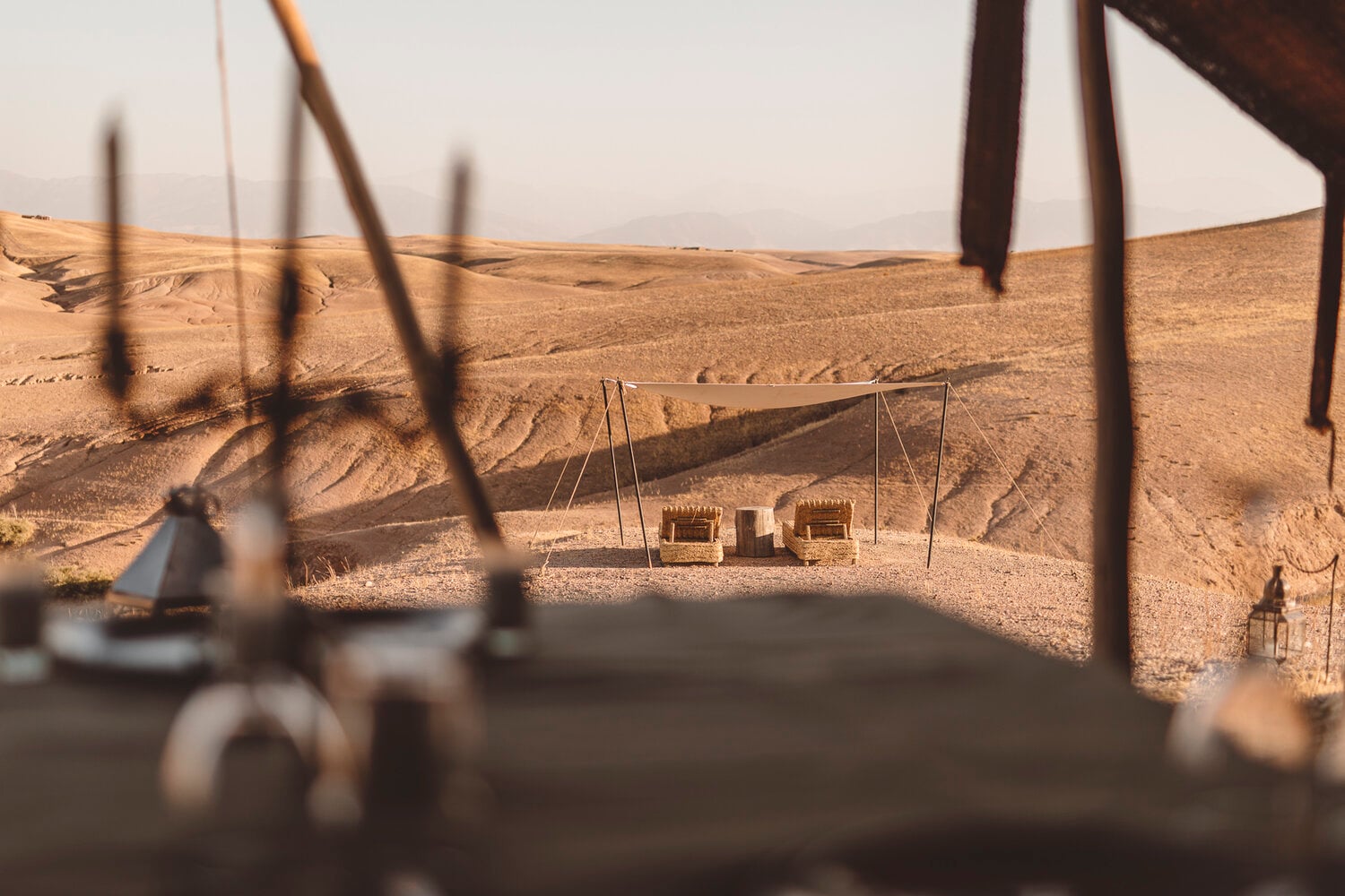 Scarabeo Camp, Africa — Desert camp with canvas pavilions and wooden structures nestled in golden sand dunes under hazy sky, viewed through blurred rope details.