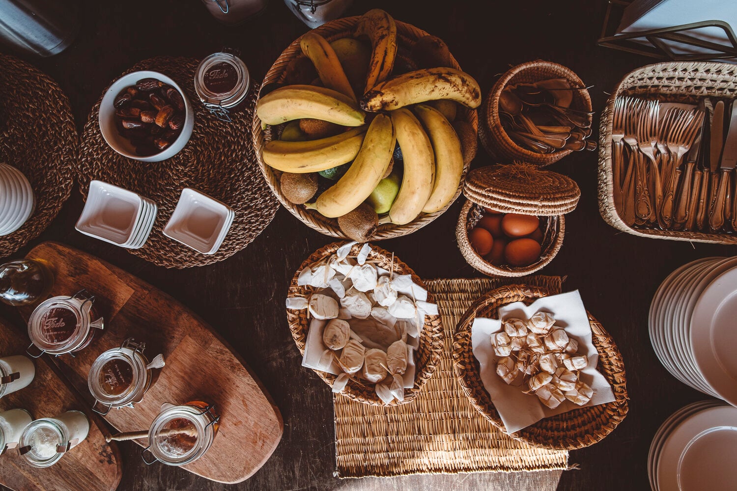 Scarabeo Camp, Africa — Overhead flatlay of woven baskets with fresh bananas, mushrooms, eggs, and artisanal tableware arranged on dark wood surface.