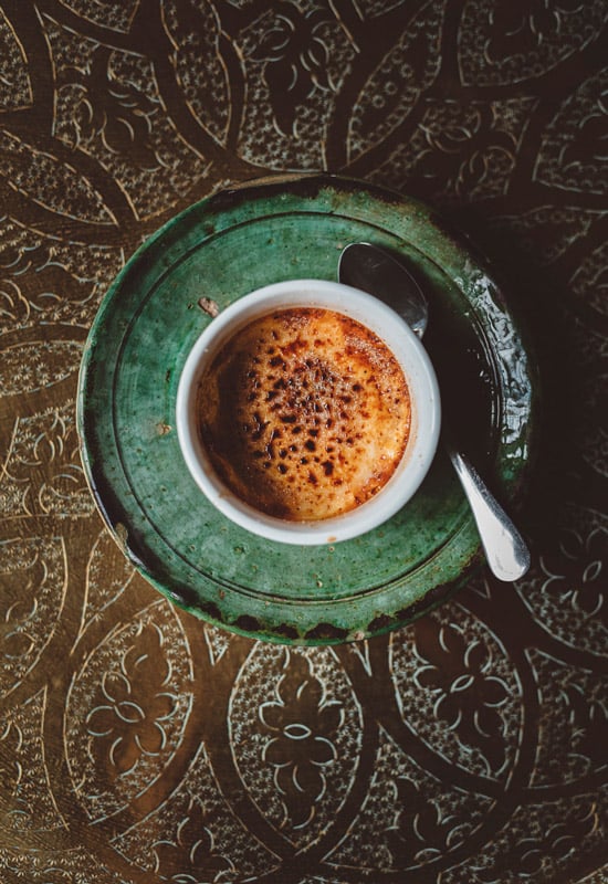Berber Lodge, Africa — Crème brûlée in white bowl on weathered green plate, caramelized sugar surface, ornate patterned textile backdrop, overhead view.
