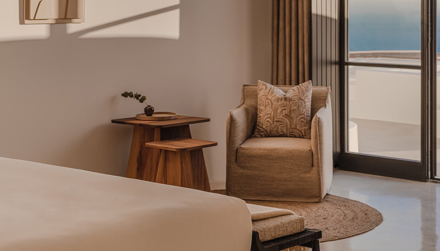 Nobu Santorini, Greece — Minimalist hotel suite with natural linen armchair, wooden side table, and expansive sea-view windows overlooking Aegean coastline.