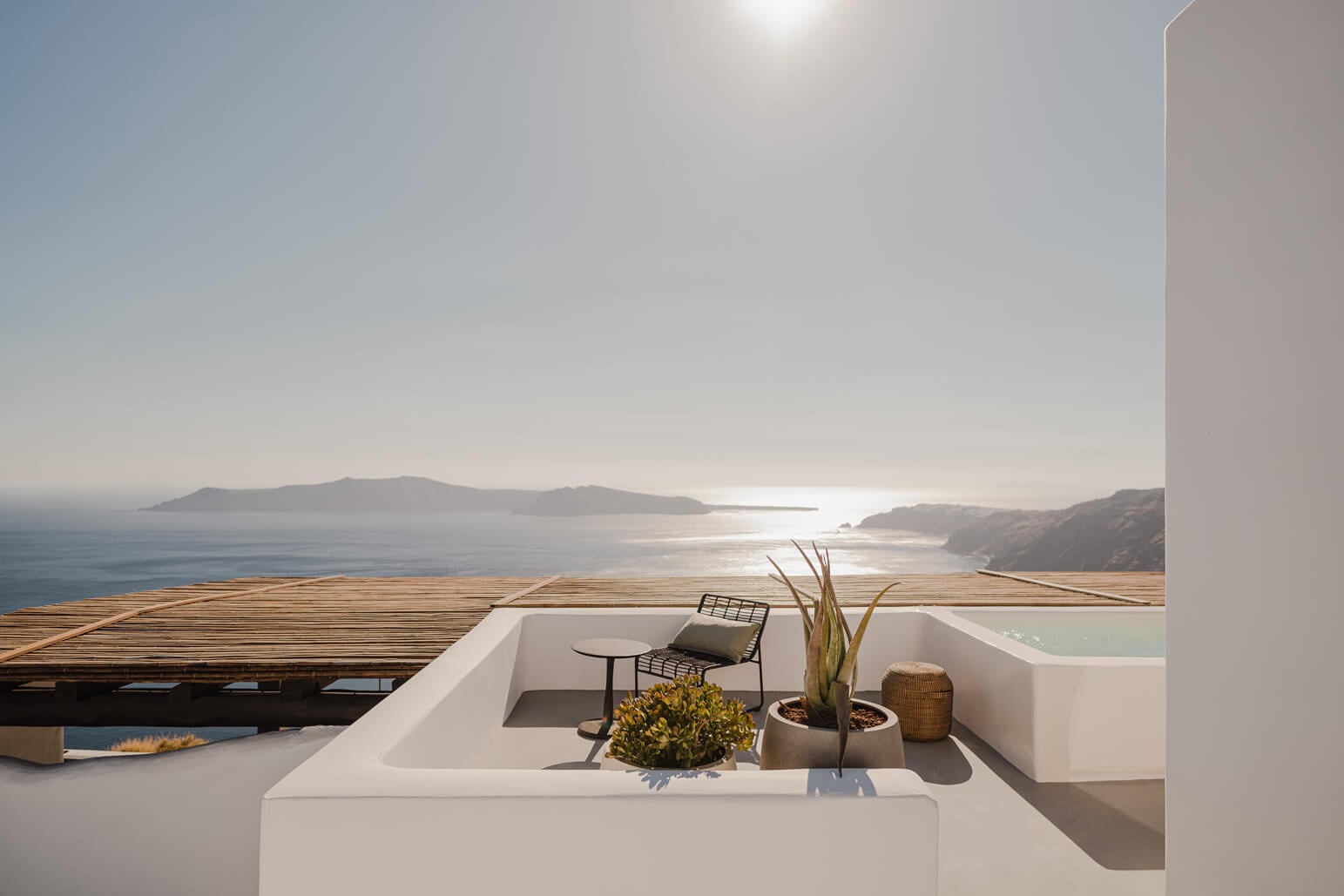 Nobu Santorini, Greece — Whitewashed terrace with wooden deck, modern seating, potted plants overlooking Aegean Sea and volcanic islands in soft daylight.