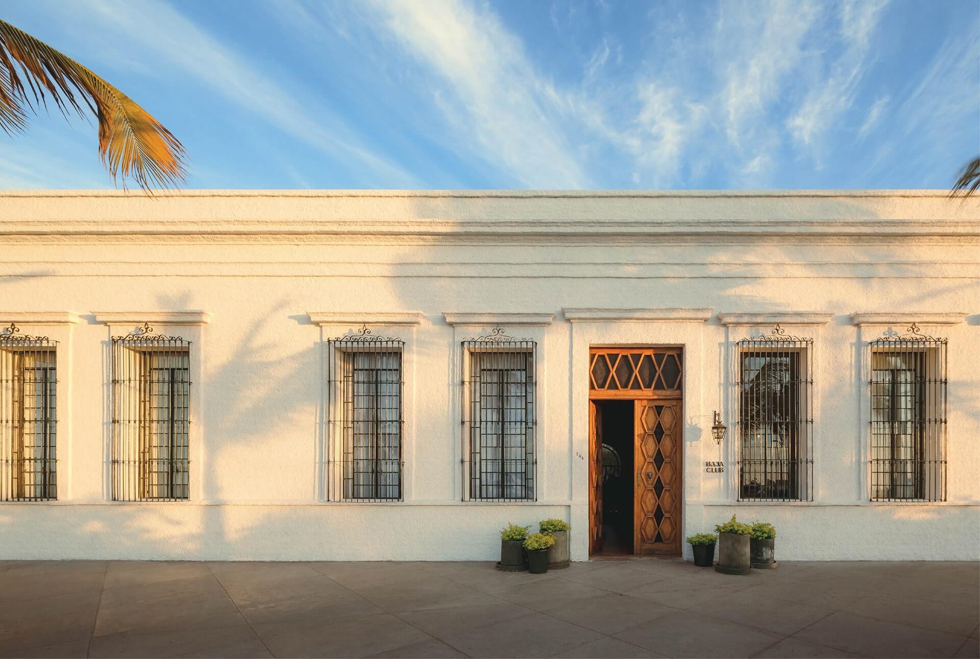 Baja Club, Mexico — Cream-colored colonial facade with iron-barred windows, wooden door, and palm shadow in bright sunlight.