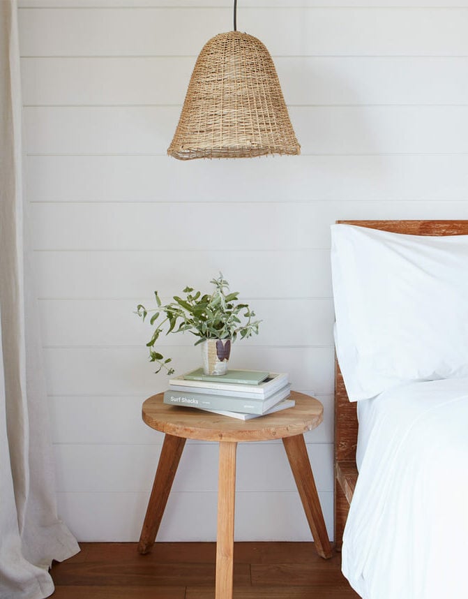 The Surfrider, North America — Coastal bedroom nightstand with woven pendant light, wooden side table, stacked books, potted plant, white linens, shiplap walls.