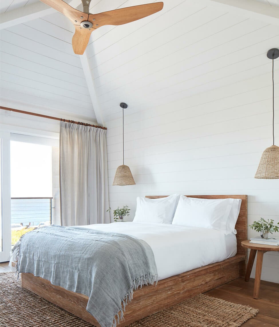 The Surfrider, North America — Coastal bedroom with shiplap walls, wooden beam ceiling fan, woven pendant lights, and ocean-view balcony access.
