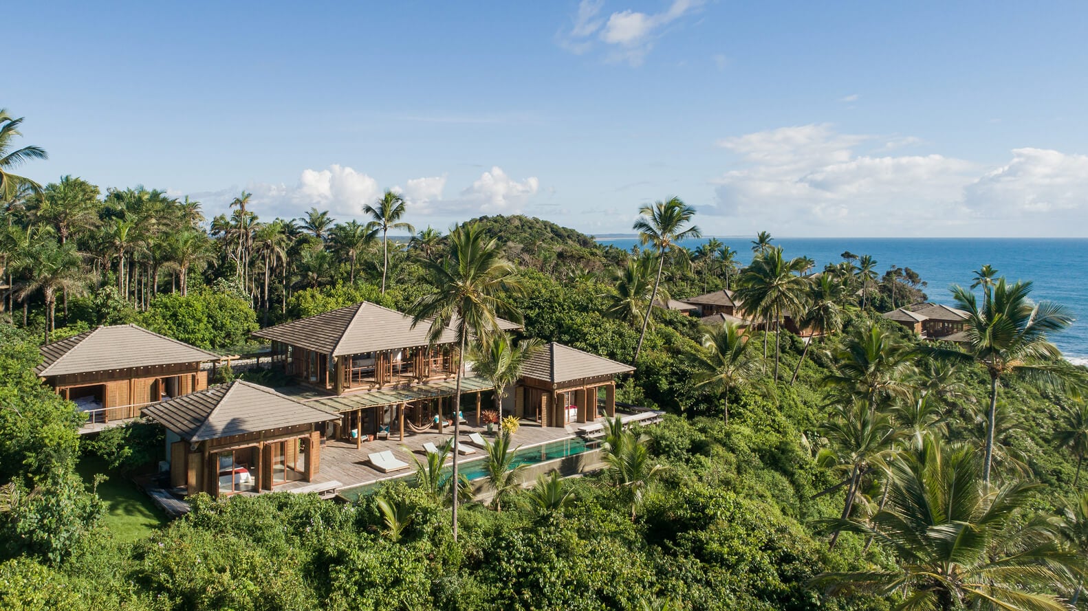 Barracuda Hotel & Villas, South America — Teak pavilions with gray metal roofing nestle in tropical hillside vegetation overlooking turquoise ocean, featuring infinity pool and palm-lined coastal architecture.